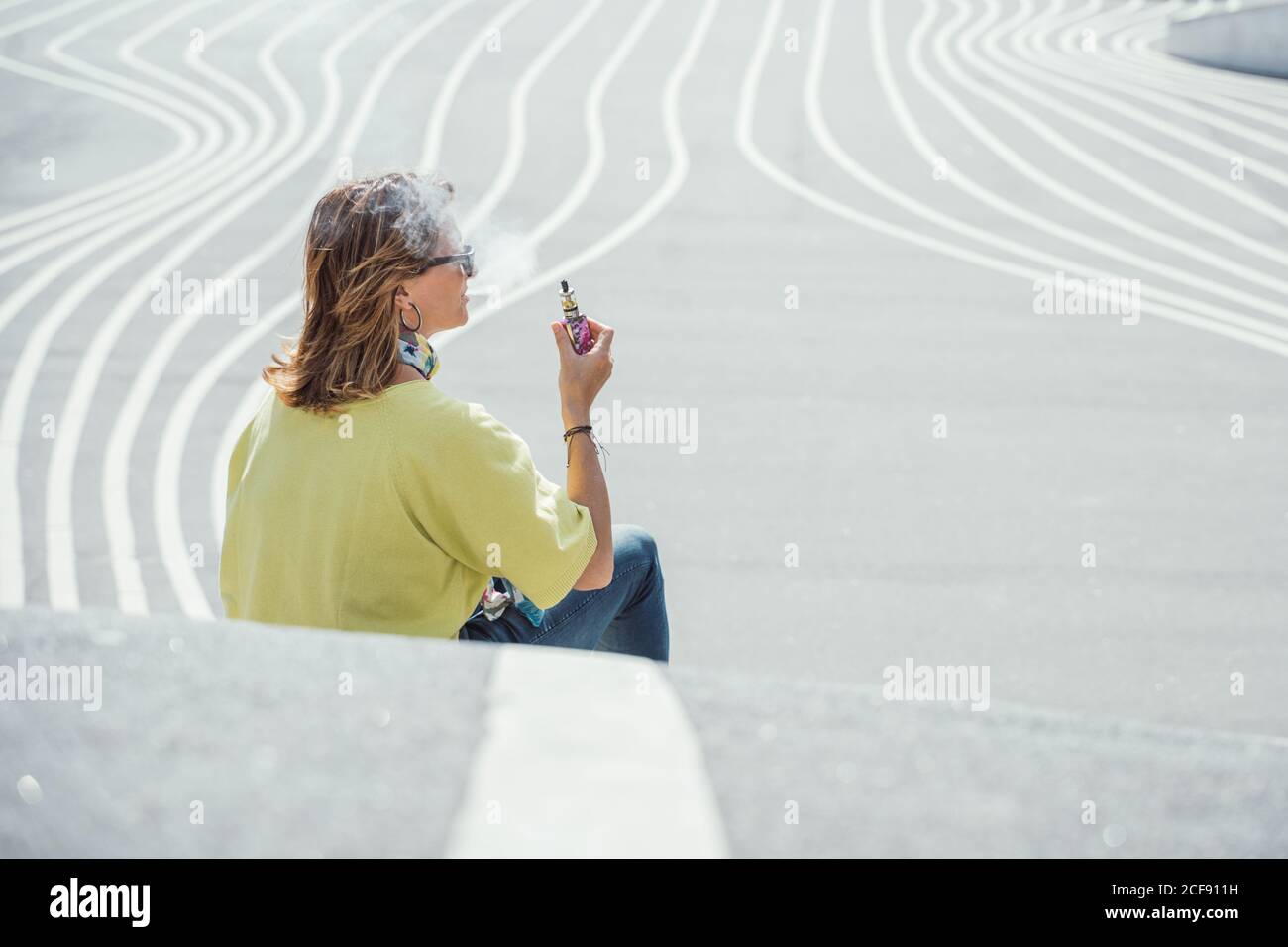 Side view of adult chilling Woman in sunglasses and colorful outfit exhaling steam while sitting on twisting asphalt road Stock Photo