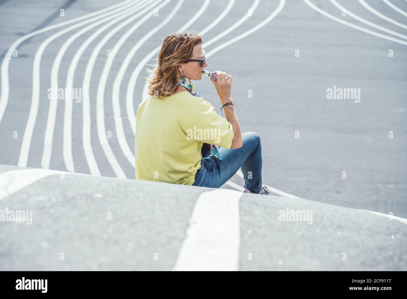 Side view of adult chilling Woman in sunglasses and colorful outfit exhaling steam while sitting on twisting asphalt road Stock Photo