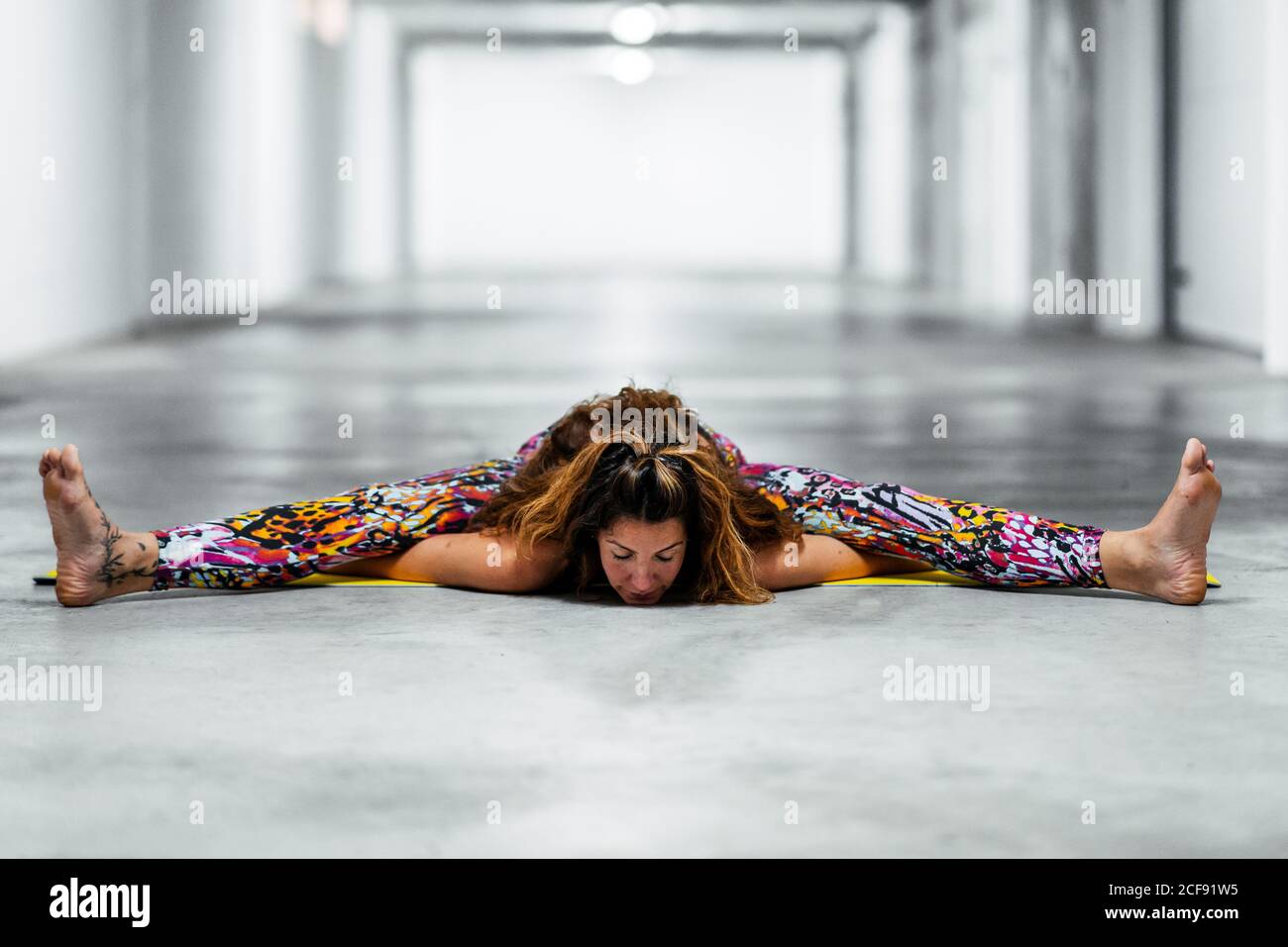 Full body young female in colorful sportswear performing Wide Angle Seated Forward Bend while practicing yoga in light garage building Stock Photo