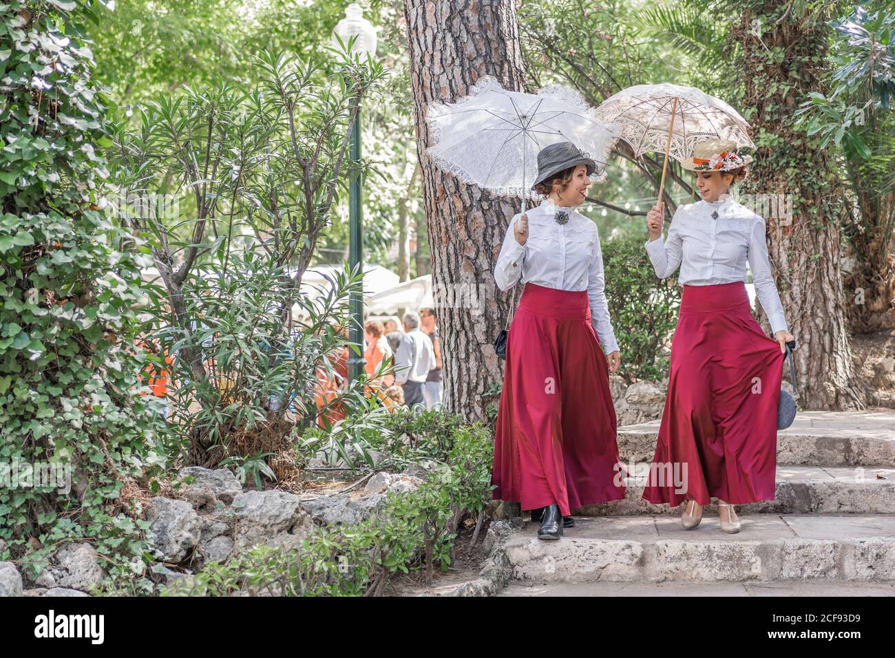 Full body ladies in retro dresses and with lace parasols smiling and talking with each other while walking down steps in park Stock Photo