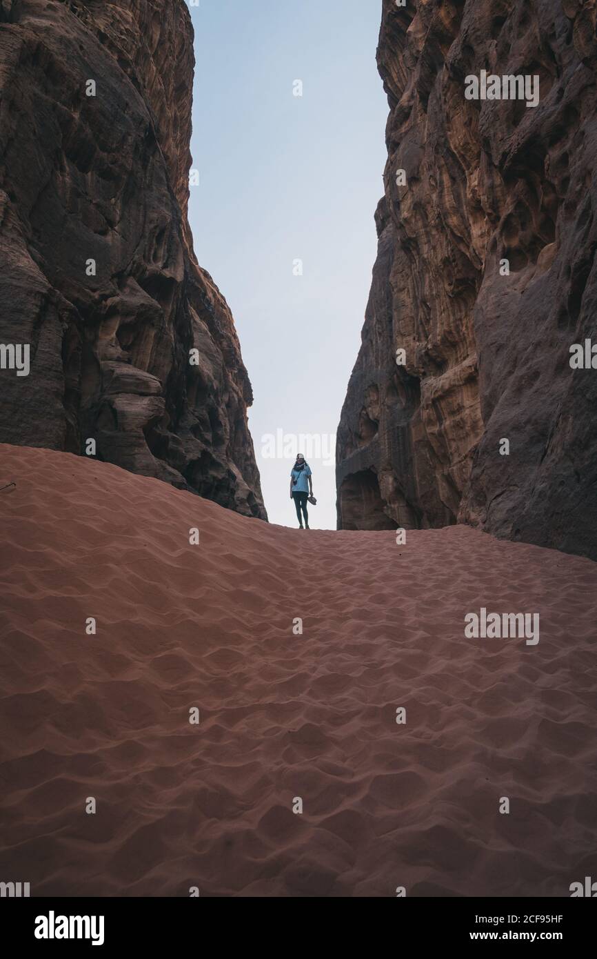 anonymous female tourist standing on sand between rough stone walls against cloudless blue sky in ravine of Wadi Rum desert in Jordan Stock Photo