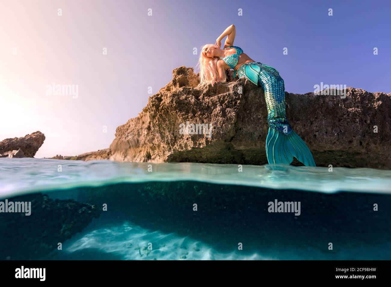 Beautiful young mermaid with blue fishtail relaxing on cliff on sunny day Stock Photo