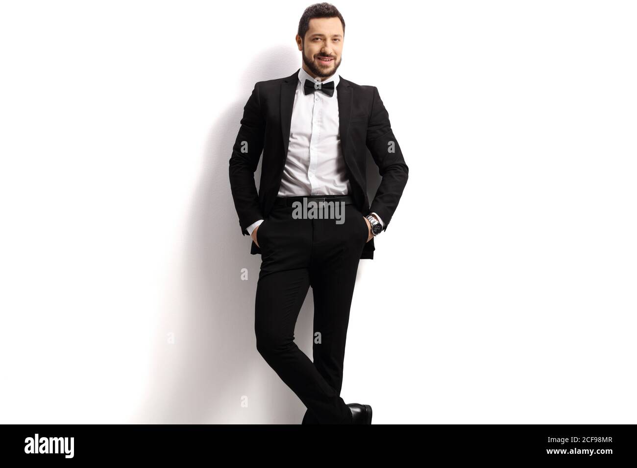 Handsome man in a black suit with bow tie leaning on a white wall Stock Photo