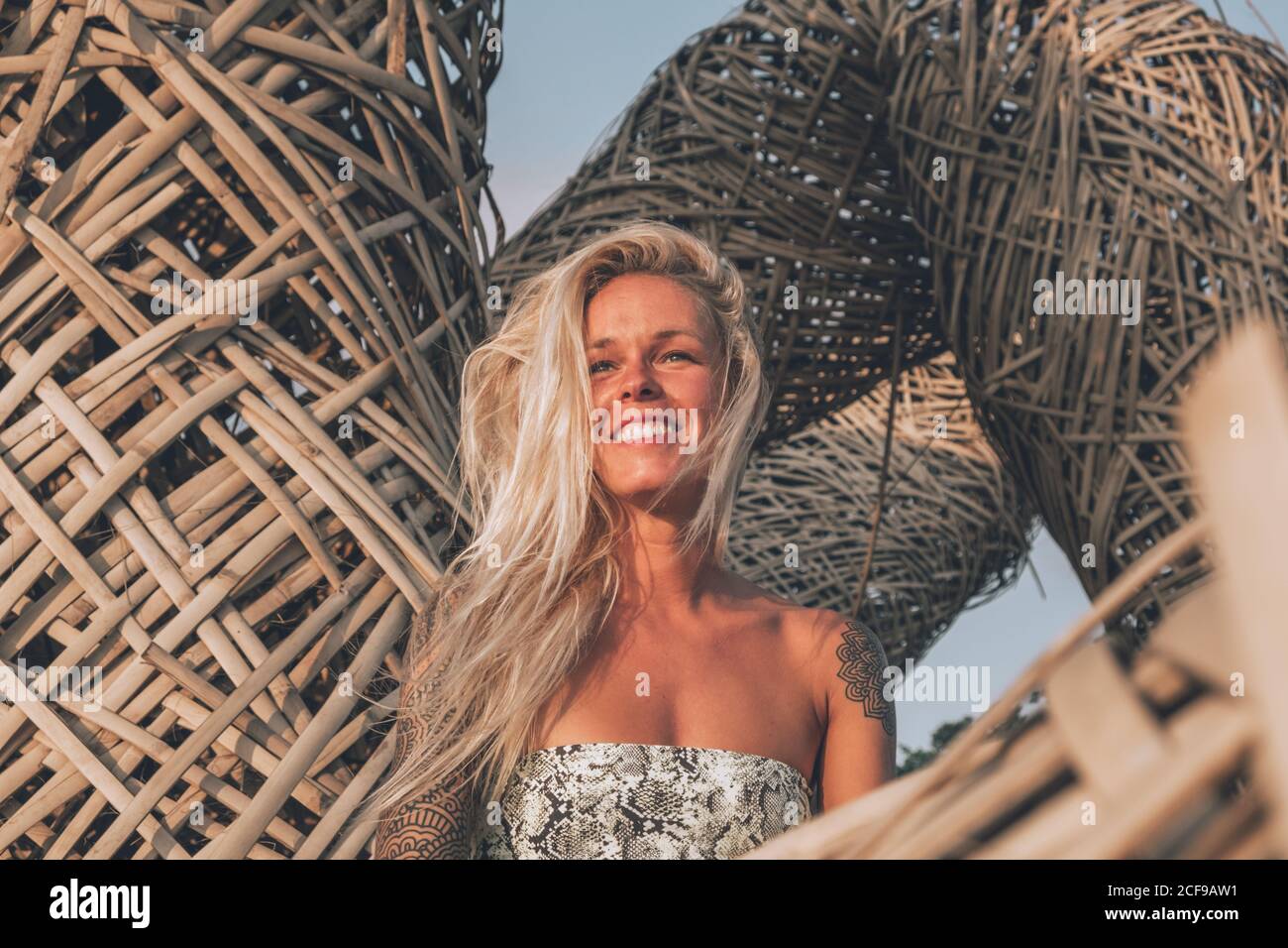 Cheerful young blonde wearing casual summer clothes with tattoos on shoulders resting on wicker seats while looking away on sunny day in summer Stock Photo