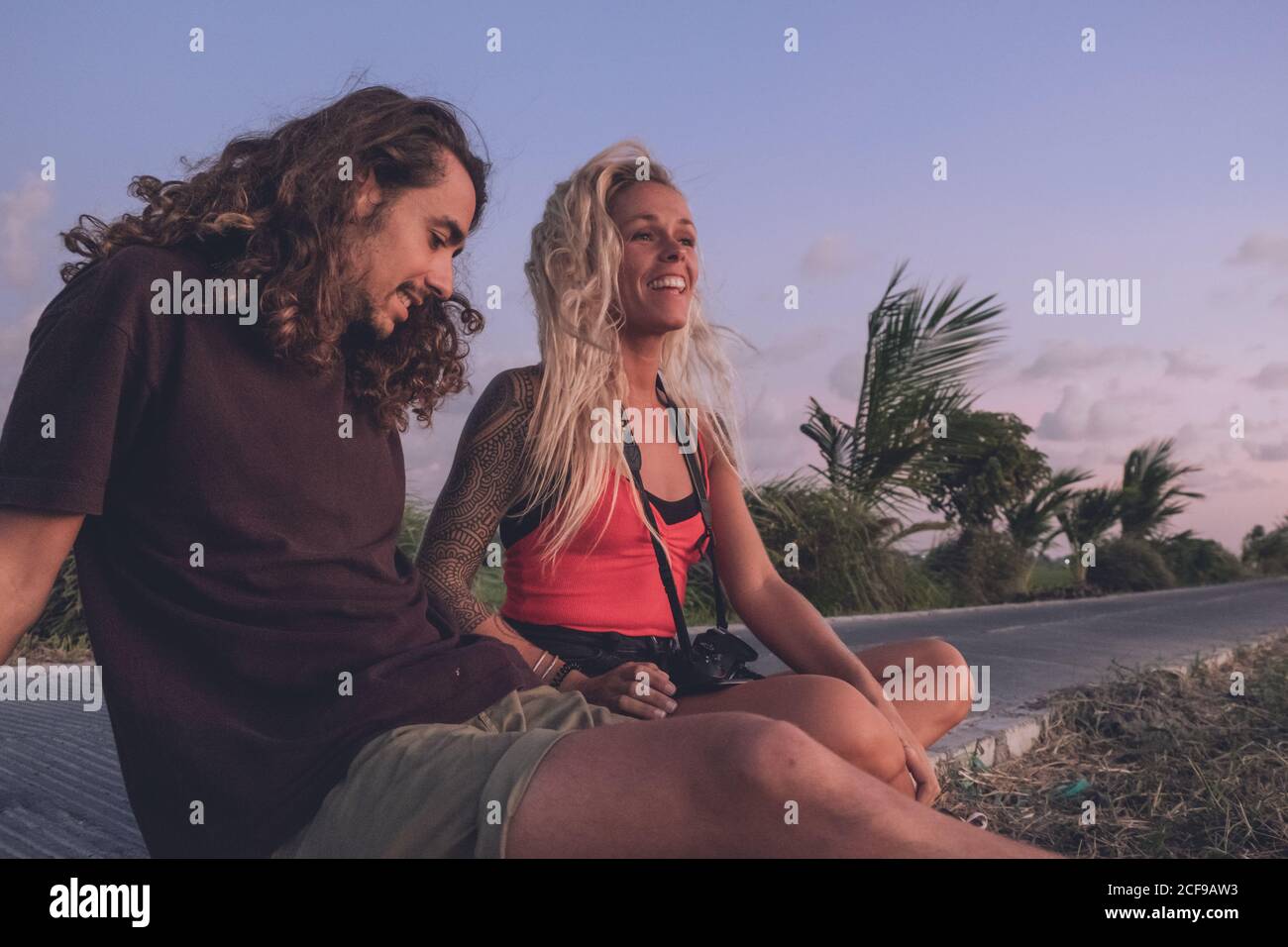 Content couple of traveling hipsters sitting on roadside in evening and watching sundown while relaxing and looking away Stock Photo