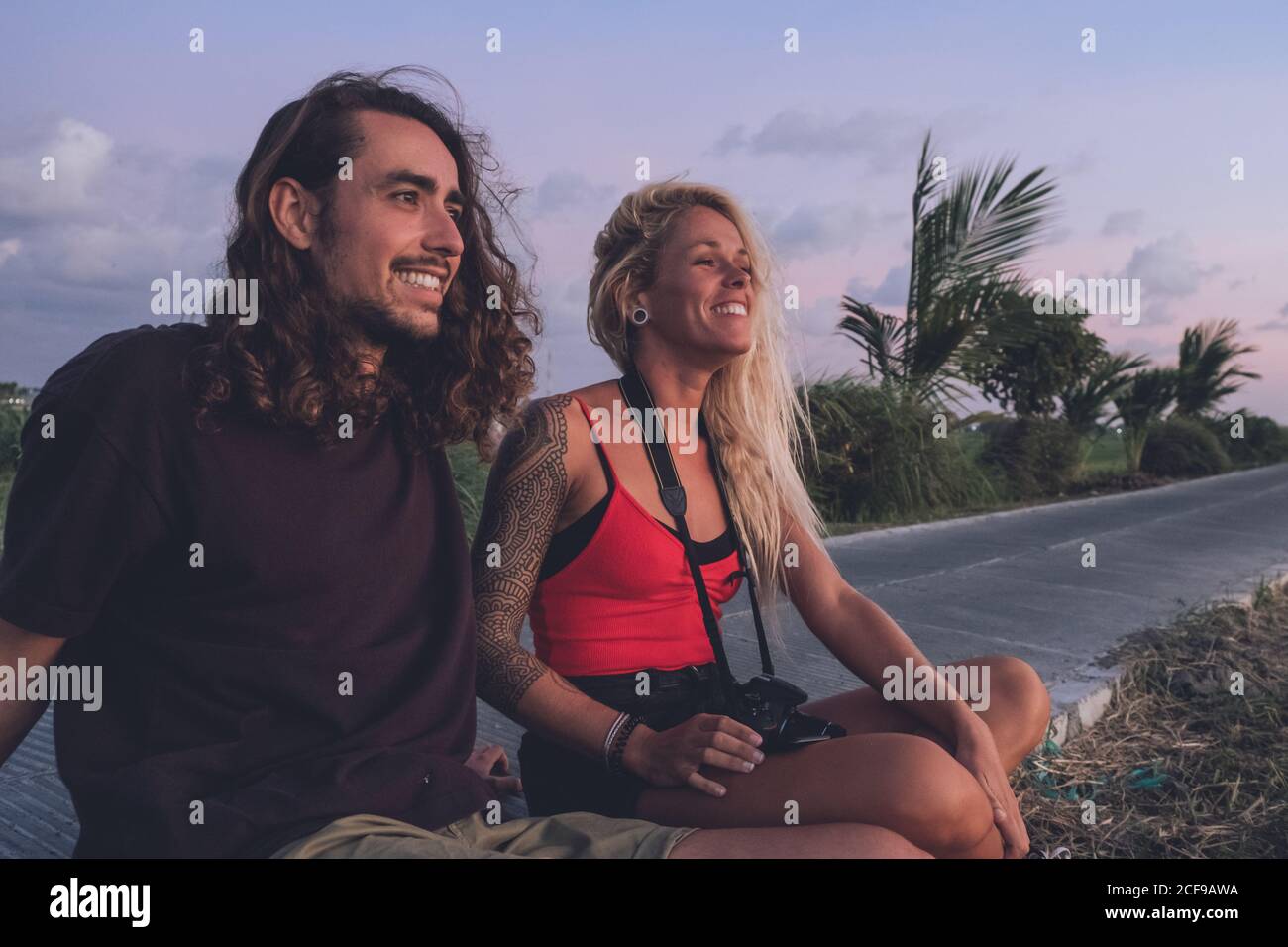 Content couple of traveling hipsters sitting on roadside in evening and watching sundown while relaxing and looking away Stock Photo