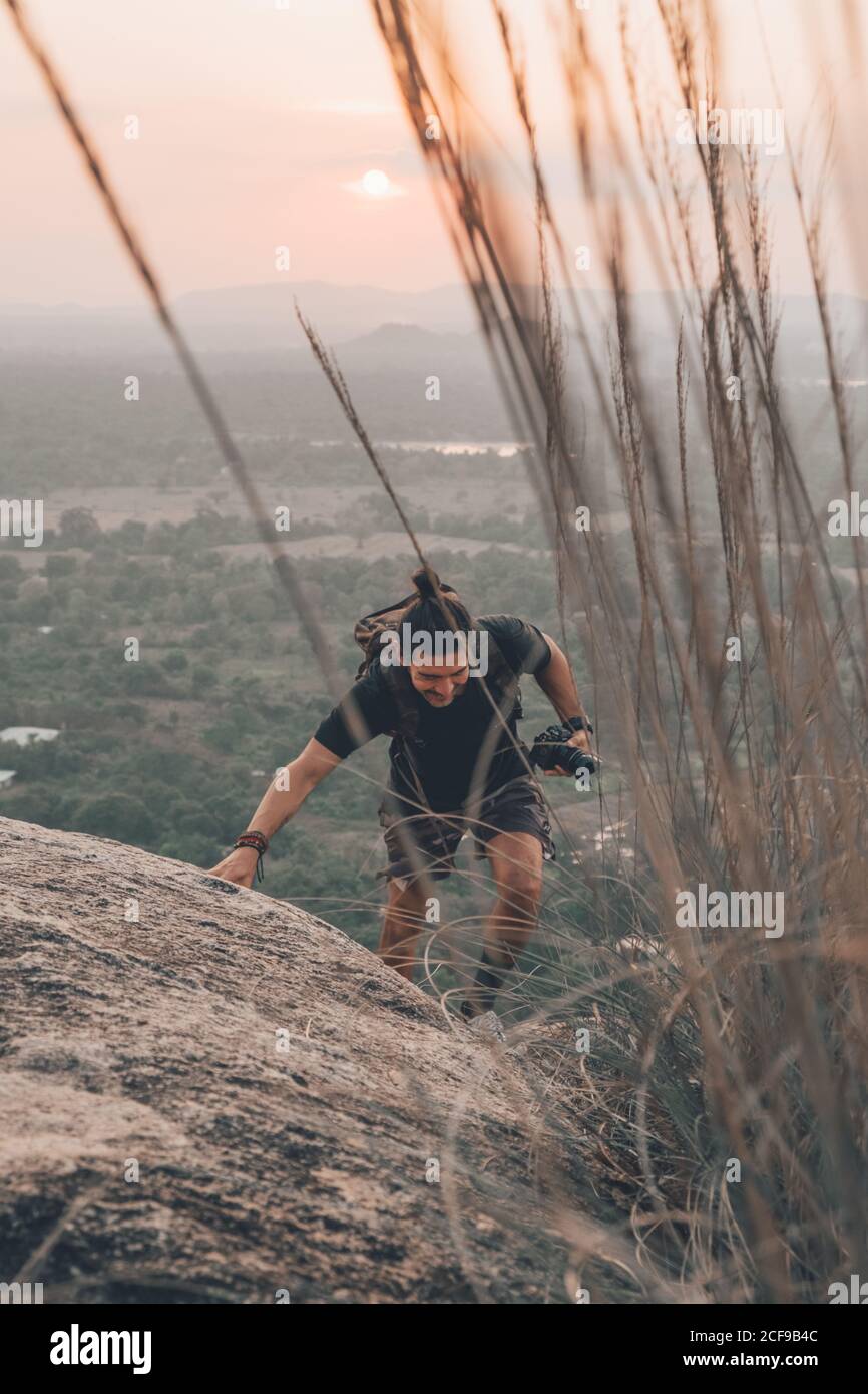 Anonymous male tourist in casual wear and backpack climbing up rocky cliff and holding  photo camera in hand while looking down on background of sunset sky Stock Photo