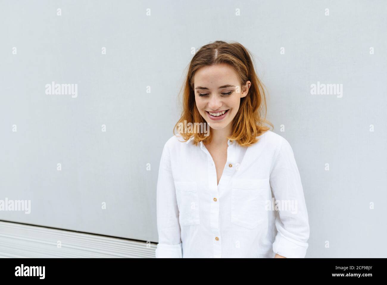Young woman standing with downcast eyes and a wide friendly smile against a white wall with copyspace outdoors Stock Photo