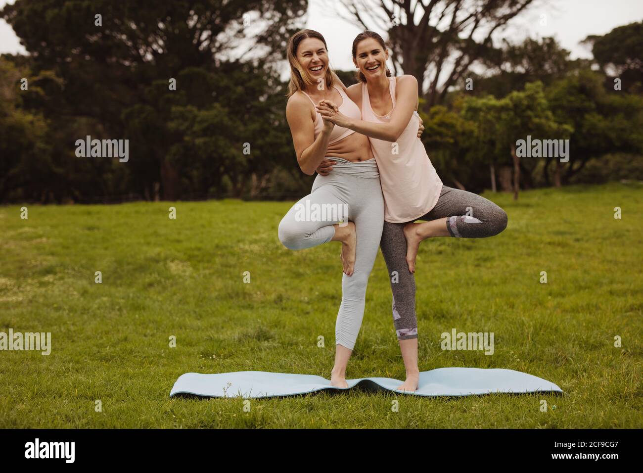 Cheerful women friends enjoying during their workout in a park. Two fitness women trying to balance while standing on one leg holding each other. Stock Photo