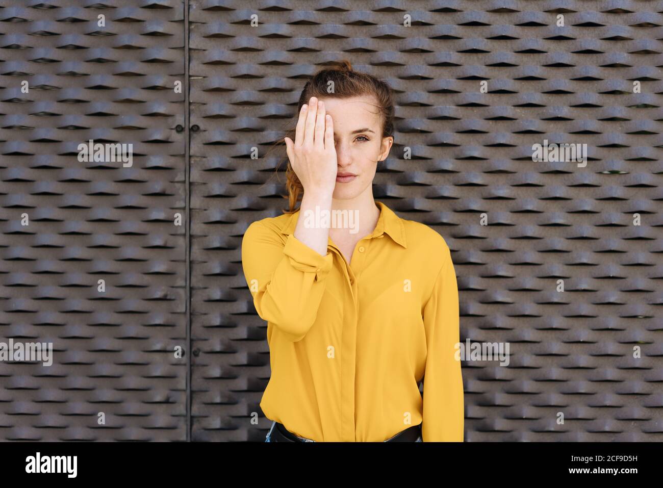Serious young woman covering one eye with her hand as she poses in front of a textured brown wall with copyspace Stock Photo