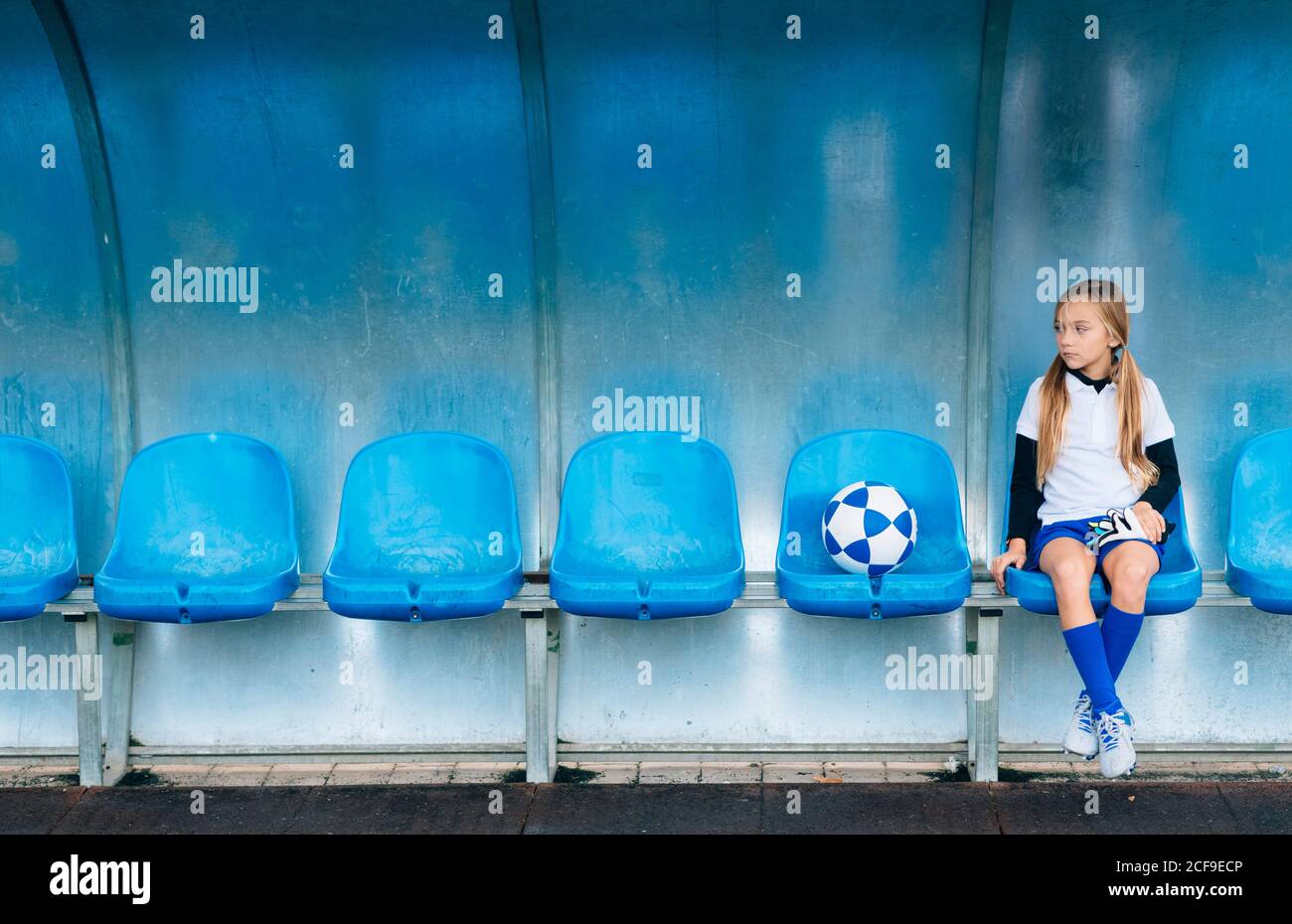 Full length frustrated preteen girl in soccer uniform sitting alone on blue plastic seat after match failure in sports club Stock Photo
