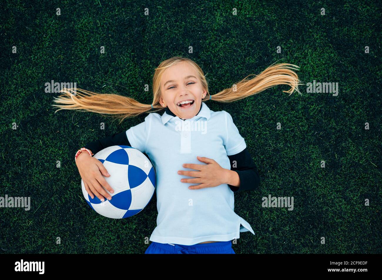From above happy preteen girl with ponytails in uniform laughing while lying with ball on green field at football stadium Stock Photo