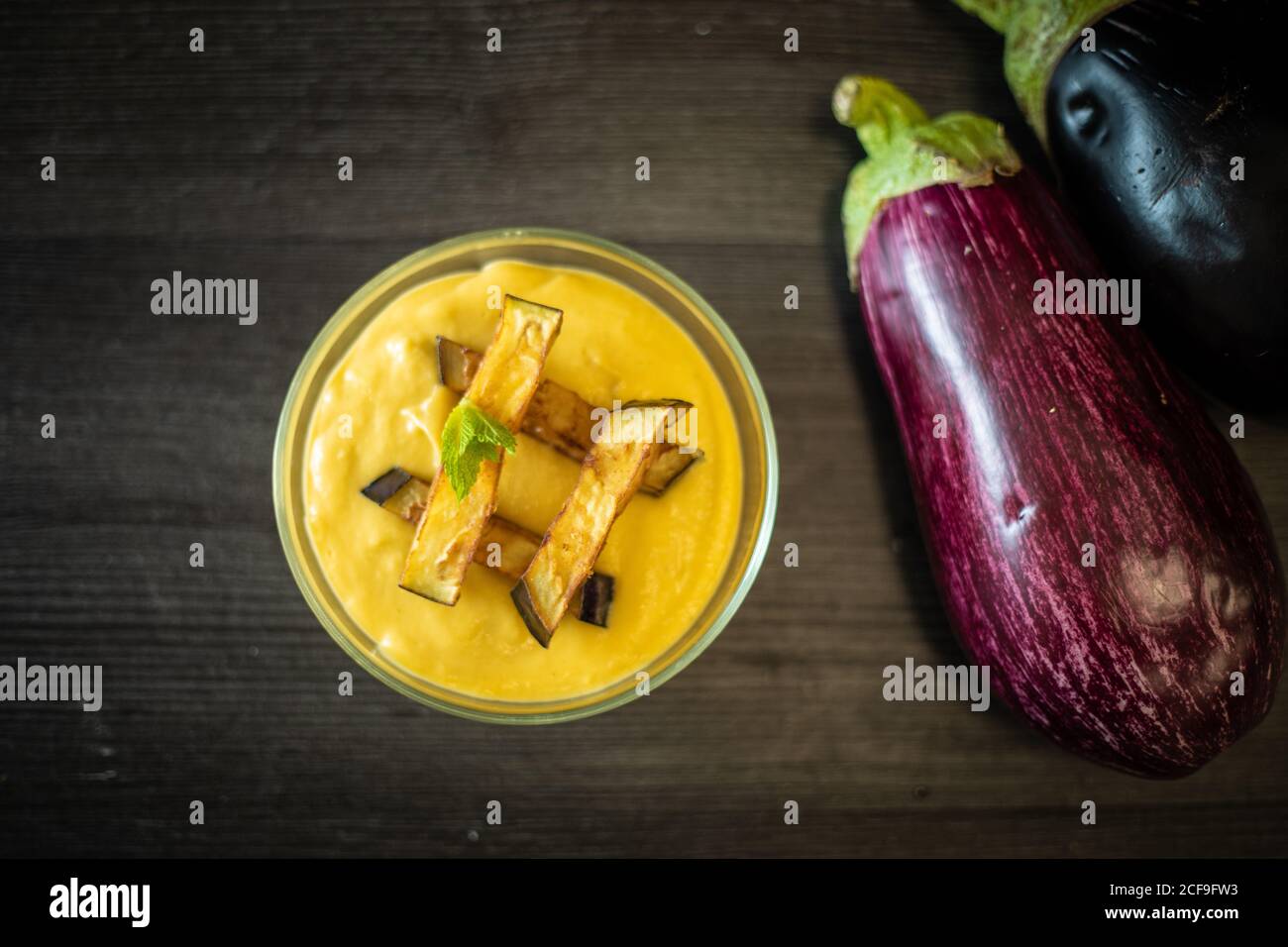 Top view of cup of tasty puree with slices of roasted eggplant and herb placed on lumber table near raw vegetables in kitchen Stock Photo
