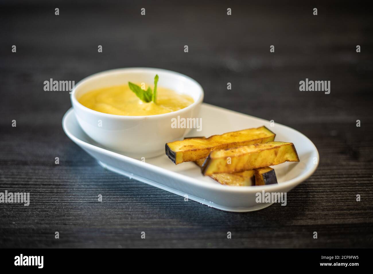 Bowl of delicious puree with herb placed on ceramic plate near pieces of fried eggplant on black wooden table in cafe Stock Photo
