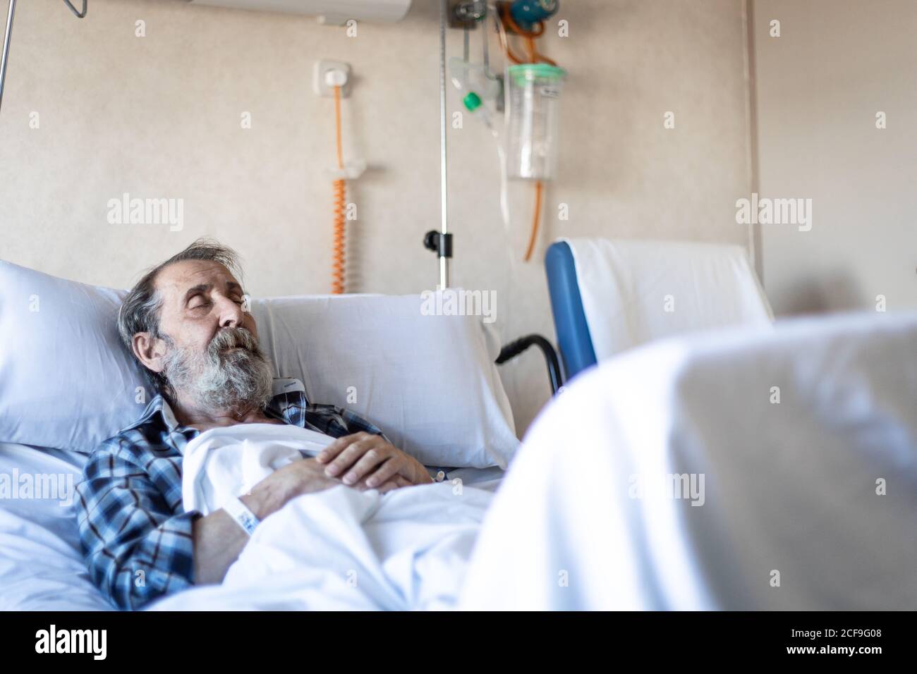 Calm aged man with beard lying under blanket on bed in hospital ward