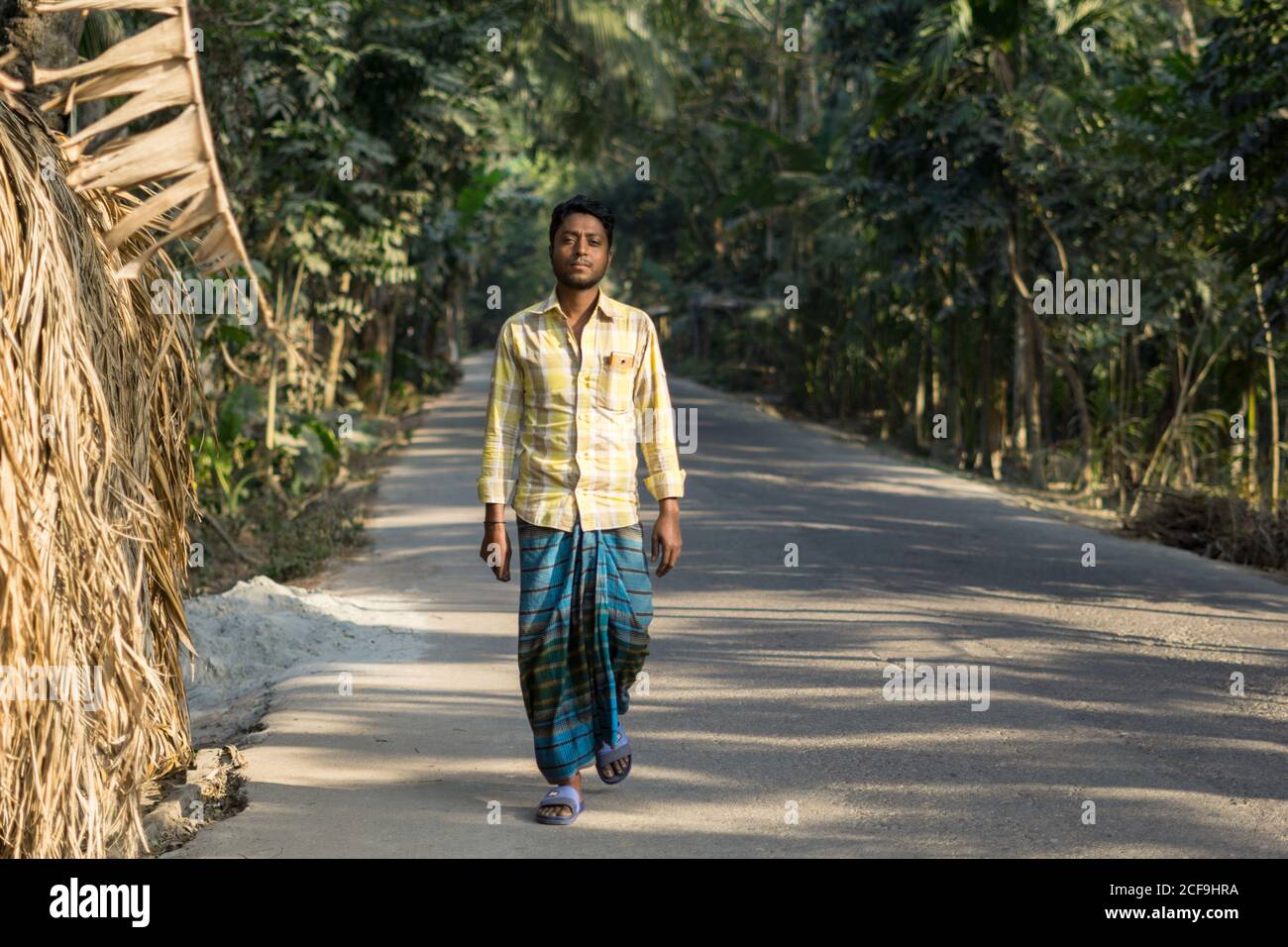 Focused ethnic male in traditional clothes looking at camera and walking on asphalt road through tropical forest in Barisal in Bangladesh Stock Photo