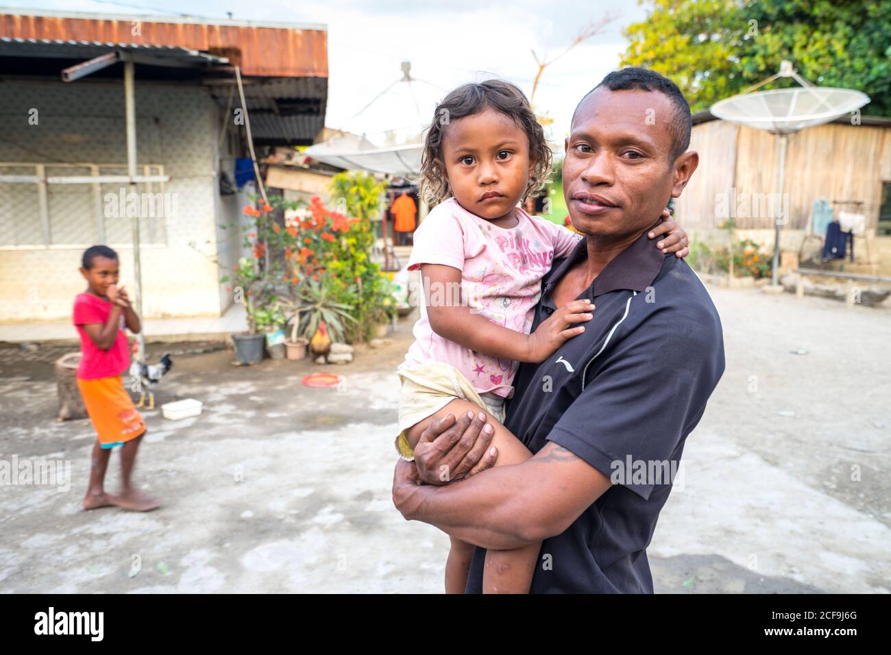 Dili, East Timor - AUGUST 08, 2018: Poor young Asian father holding little girl and looking at camera while standing on street next to house with boy in background Stock Photo