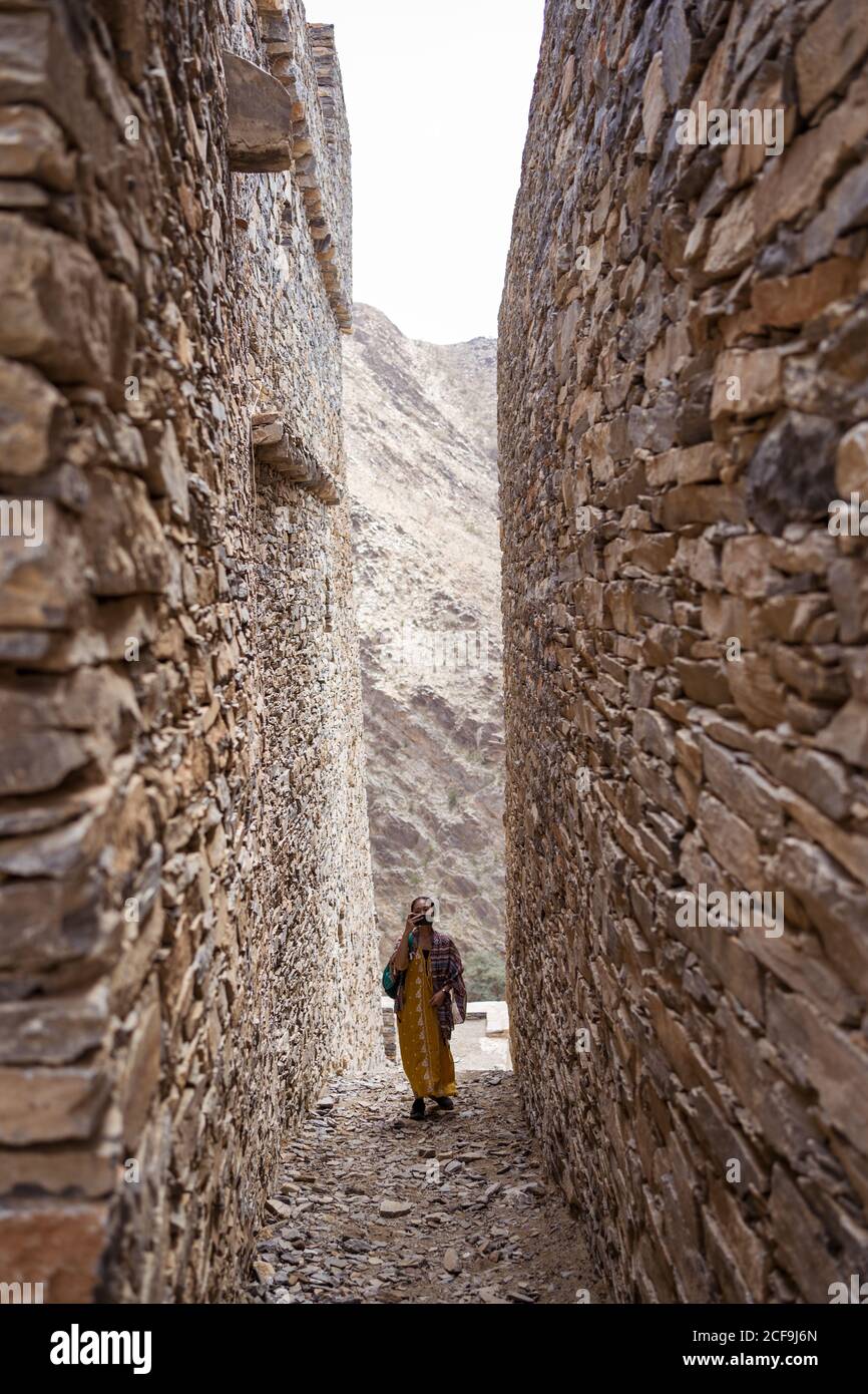 Distant Woman in colorful clothes walking between shabby ancient walls of historic buildings of Marble Village in Al Bahah locating on mountain terrain in Saudi Arabia Stock Photo