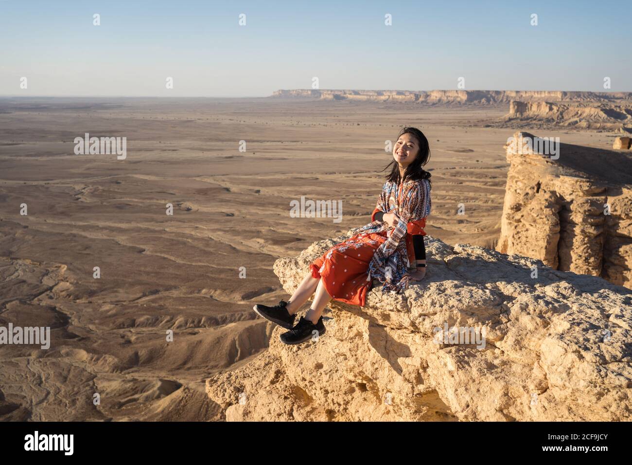 Side view of ethnic asian Woman in casual clothes sitting on cliff smiling and looking at camrea in the Edge of the World in Saudi Arabia Stock Photo