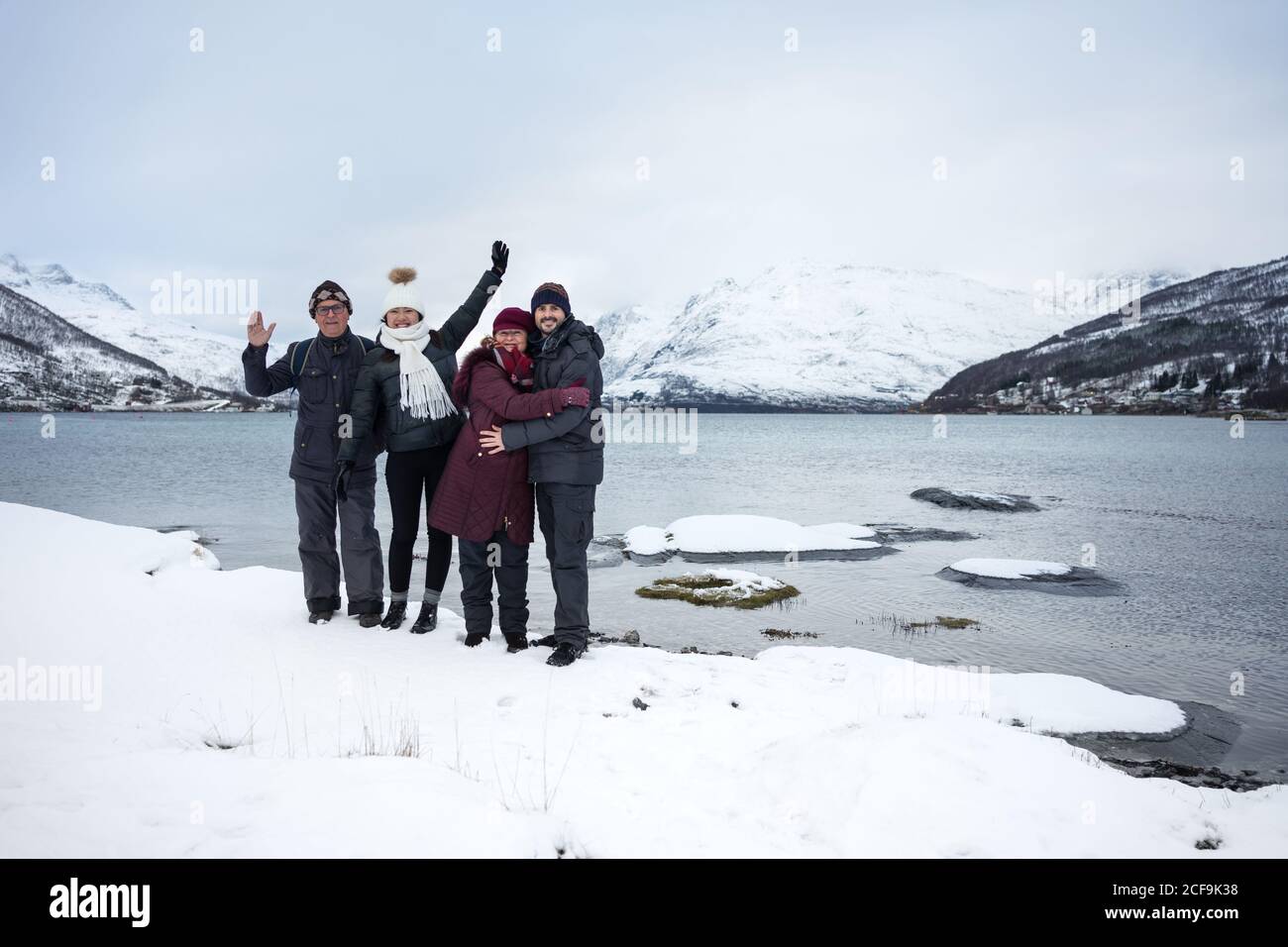 Cheerful people in warm clothes smiling at camera while standing on Kaldfjorden lake shore and waving hands against snowy hills in Norway Stock Photo