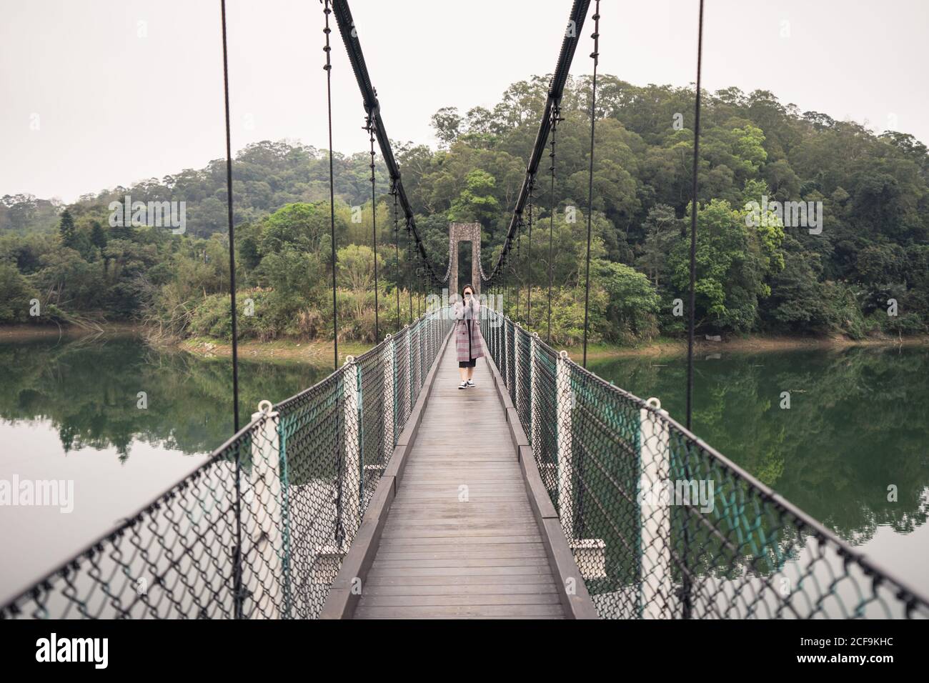 Female traveler in coat taking photo with photo camera while standing on wooden suspension bridge with metal fence over lake near green forest Stock Photo
