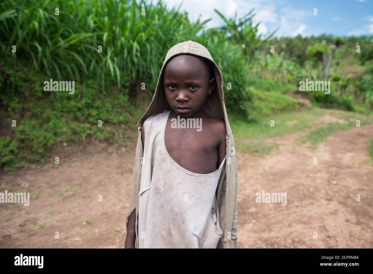 Ruanda, Africa - December 14, 2019: Miserable little African boy in dirty clothes walking on road in green rural land looking at camera Stock Photo