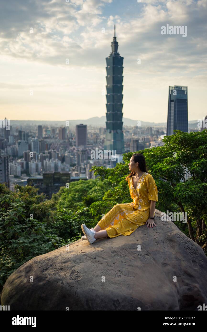 Tranquil female traveler in stylish yellow dress sitting in big rock while enjoying in views with city plants and sky on background in Scotland Stock Photo
