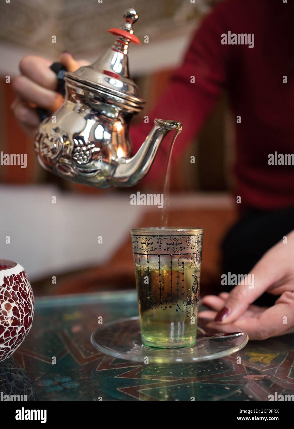 Happy ethnic female pouring fresh Arabic tea in cup while sitting on comfortable couch in small ornamental room Stock Photo
