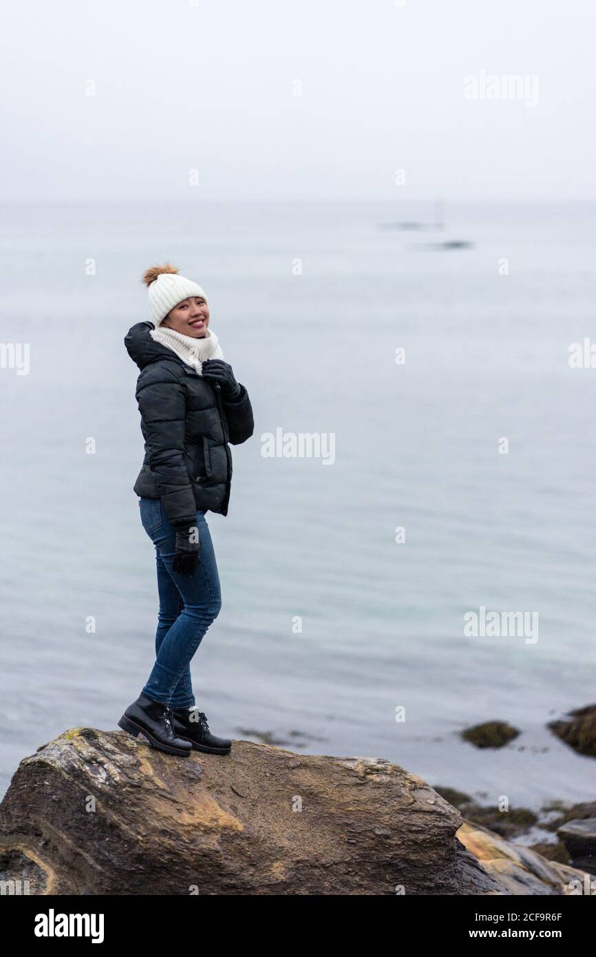 Side view of Woman in warm clothes looking at camera on small rocky ledge and enjoying beauty of northern landscape in Tromso, Norway Stock Photo