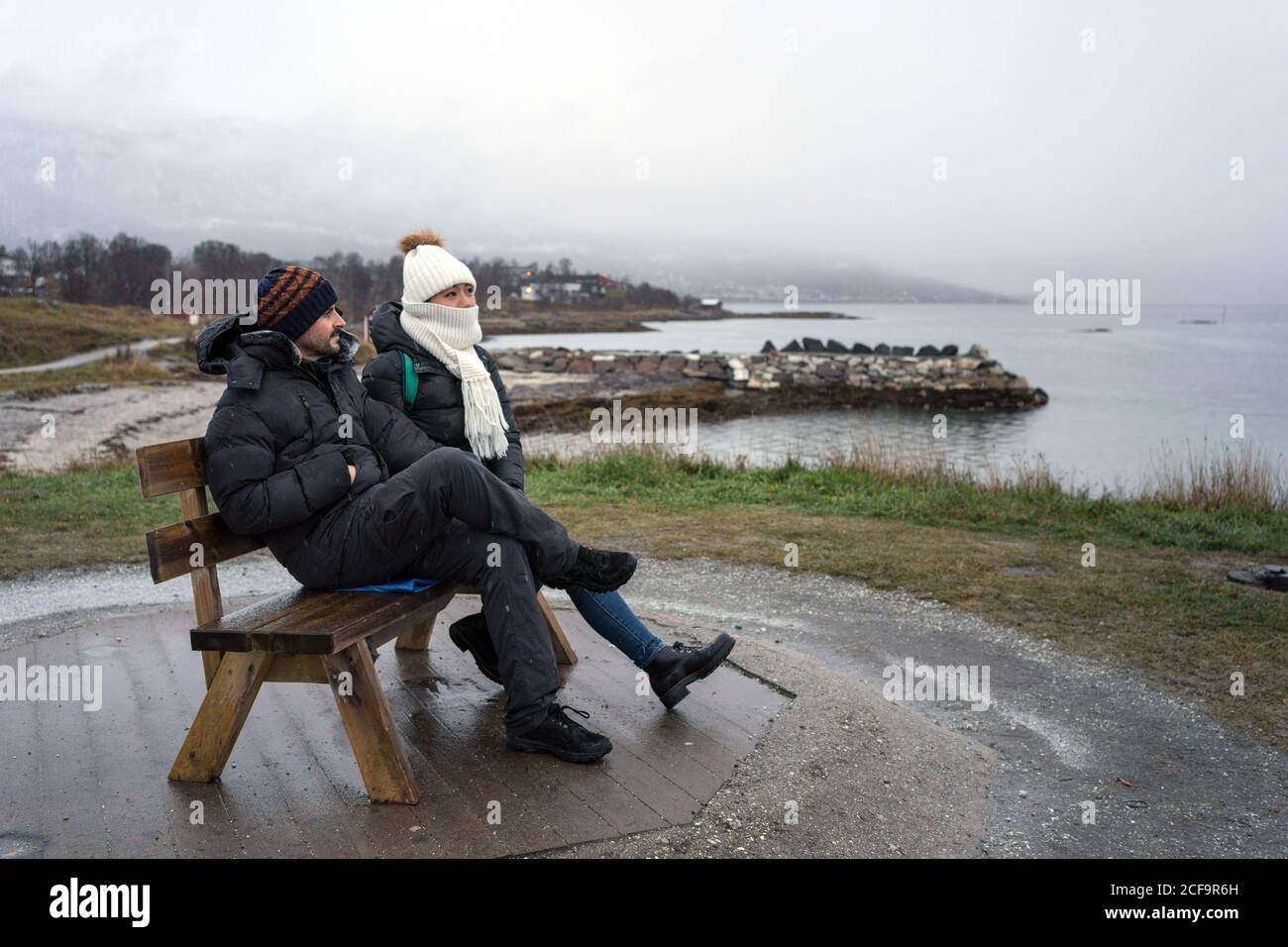 Frozen young friends in warm clothes sitting on wooden bench of ...