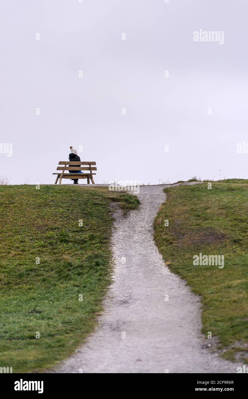 Back view of anonymous lonely female tourist resting on wooden bench in rainy weather on elevated location near sea in Tromso, Norway Stock Photo