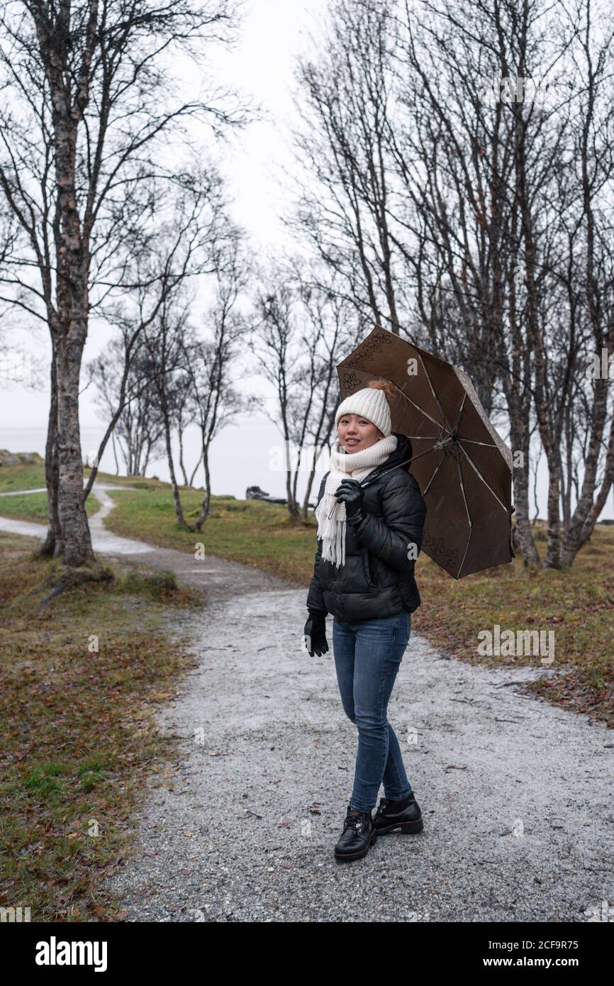 Content tourist in warm clothes with umbrella during stroll on rainy weather in park zone of Tromso, Norway looking at camera Stock Photo