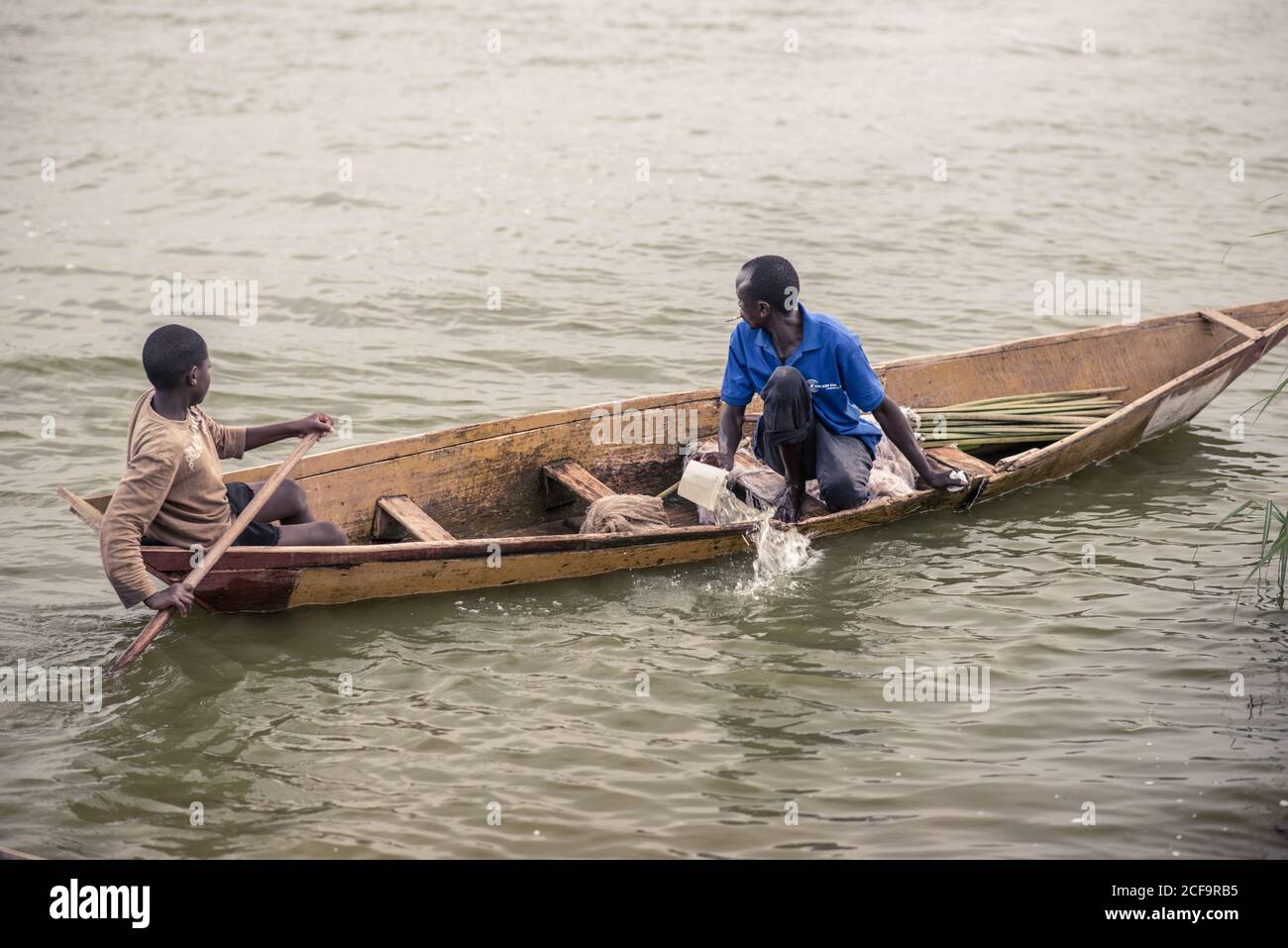 Uganda - November, 26 2016: Male teenager rowing while adult man bailing out water from old boat and looking away during ride on dirty river Stock Photo