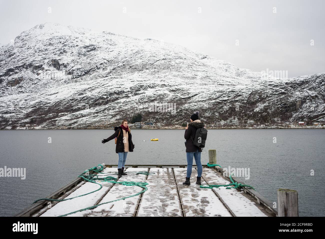 Ersfjordbotn, Norway - October 25, 2019: Tourists in warm clothes having conversation while standing on wooden pier and enjoying view of tranquil lake and hillside covered with snow in quiet cold overcast weather Stock Photo