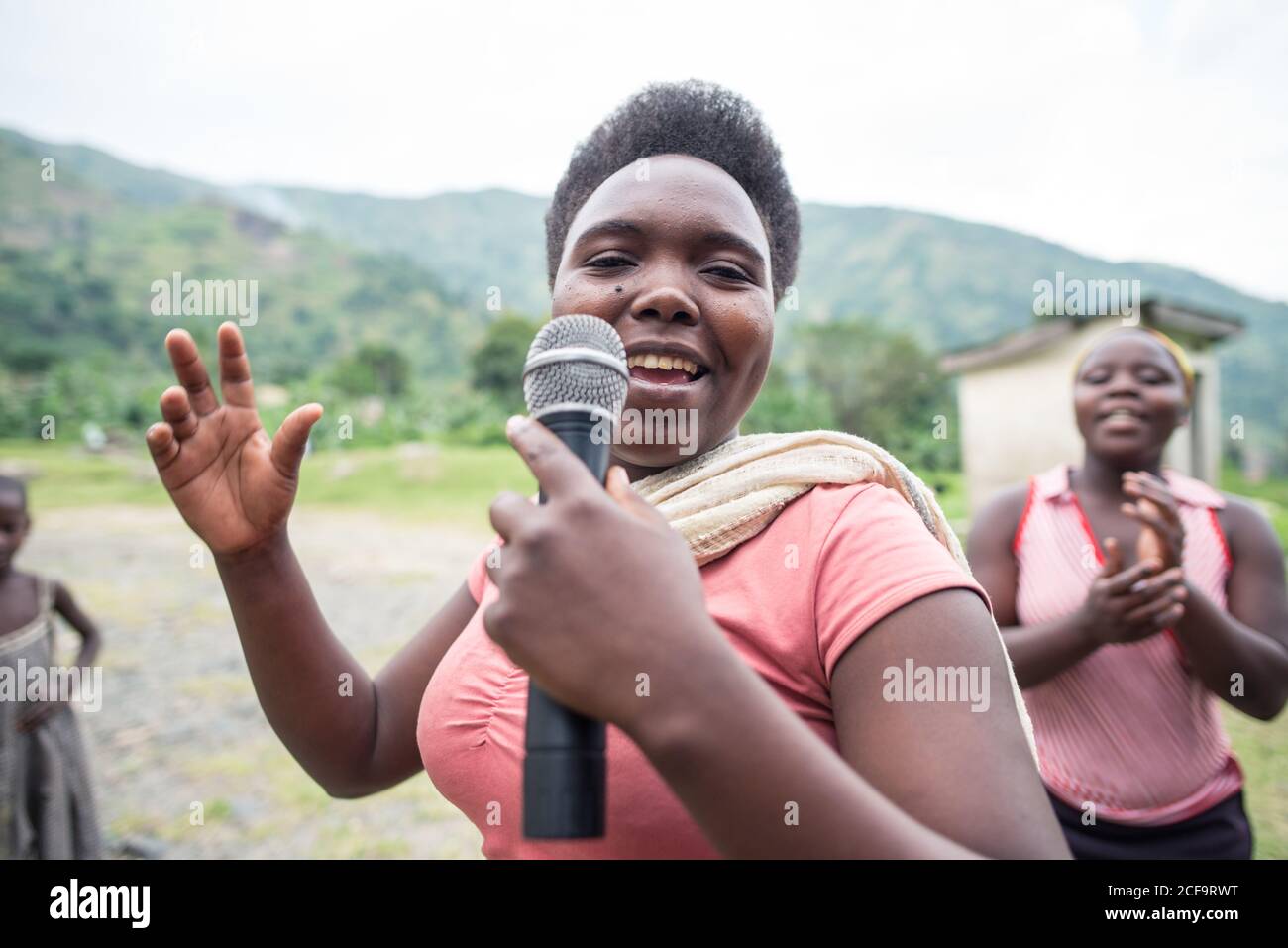 Uganda - November 26, 2016: Cheerful African female in pink shirt holding microphone and singing song while excited audience listening with interest and clapping standing against blurred green hill and small building in village Stock Photo