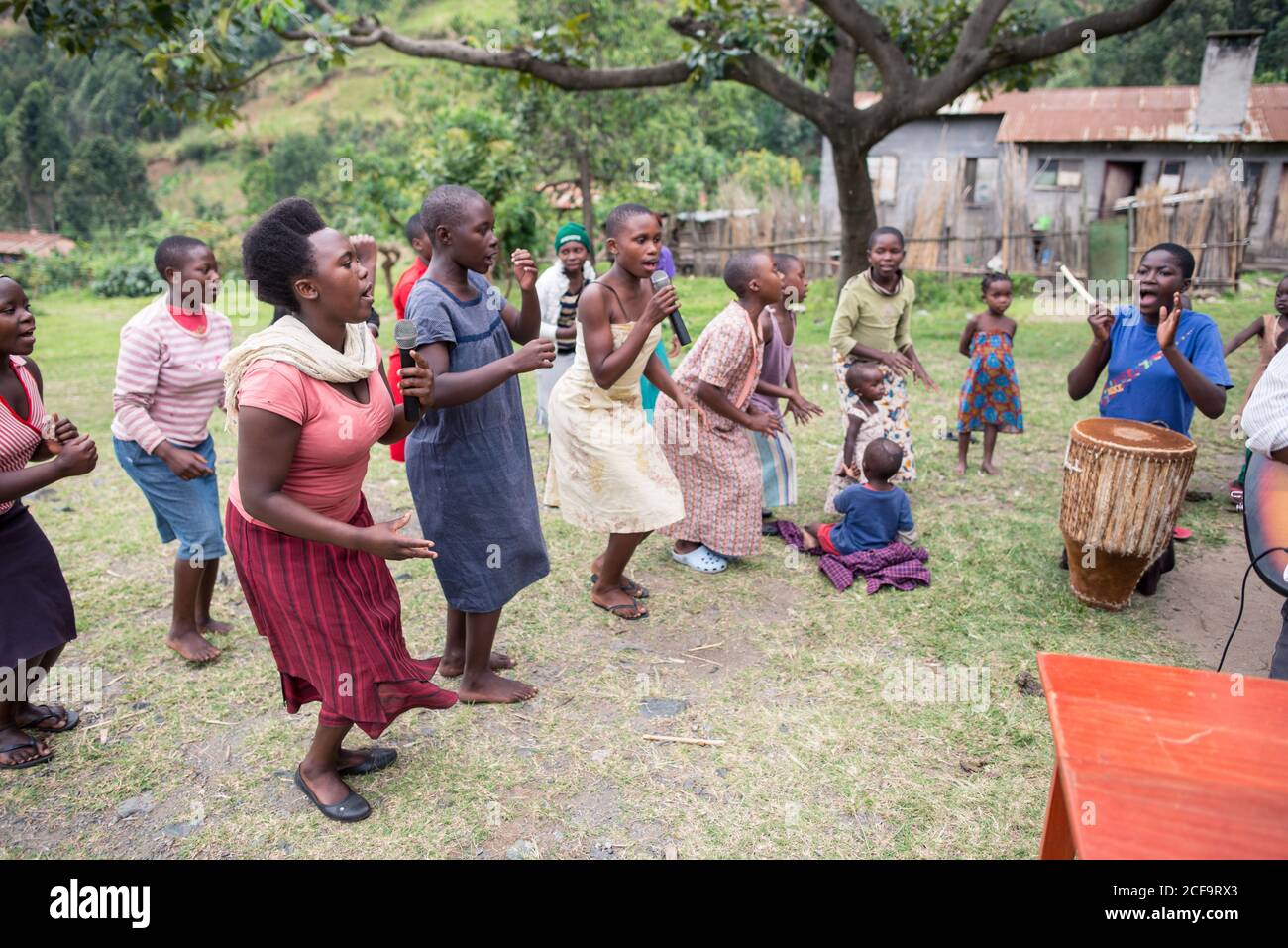 Uganda - November 26, 2016: Cheerful African female in pink shirt holding microphone and singing song while excited audience listening with interest and clapping standing against blurred green hill and small building in village Stock Photo