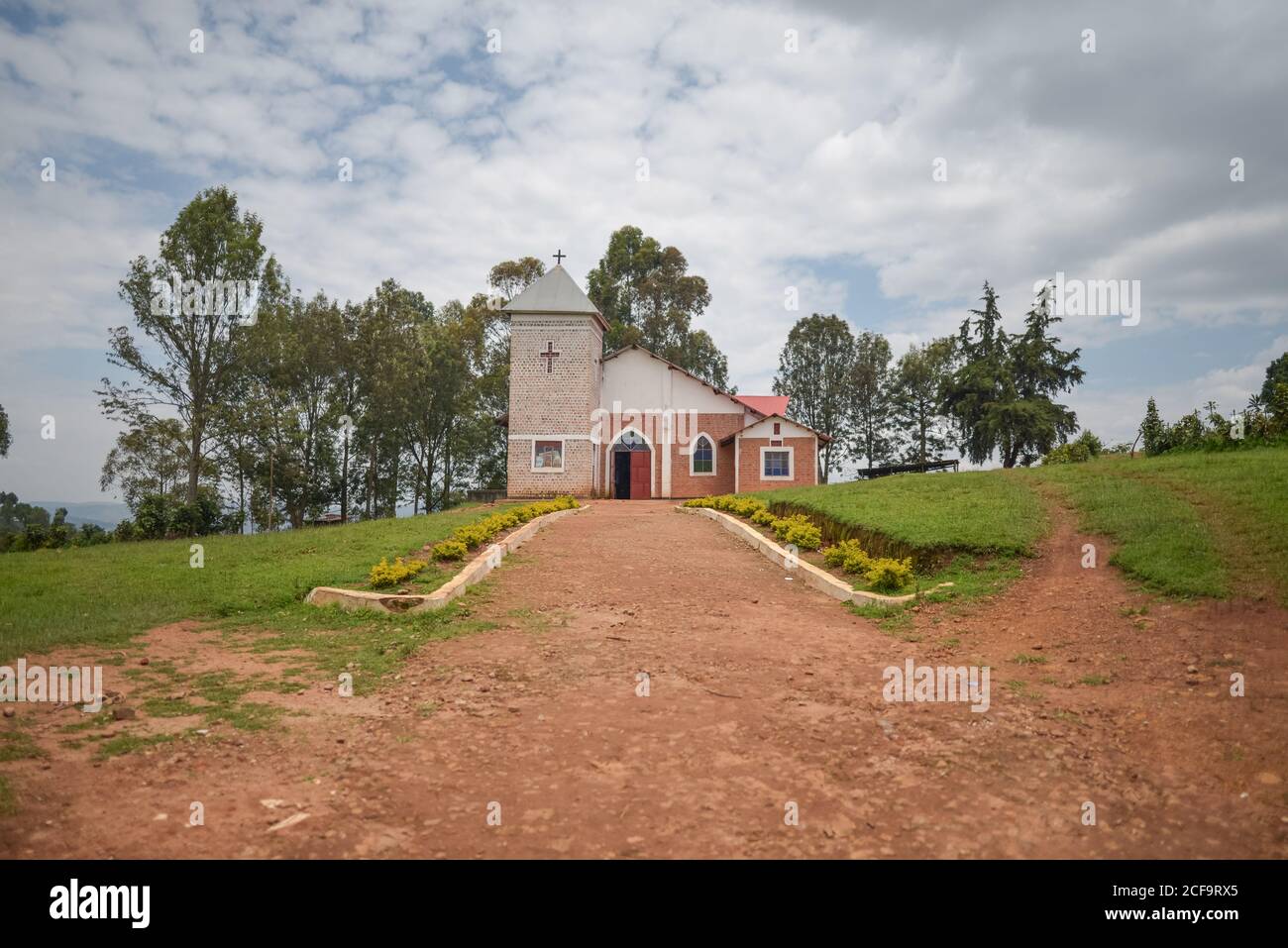 Uganda - November 26, 2016: Empty dirt footpath crossing cozy yard and leading to small white and brown brick chapel located on grassy hillside against tall green trees under blue sky with lush clouds Stock Photo
