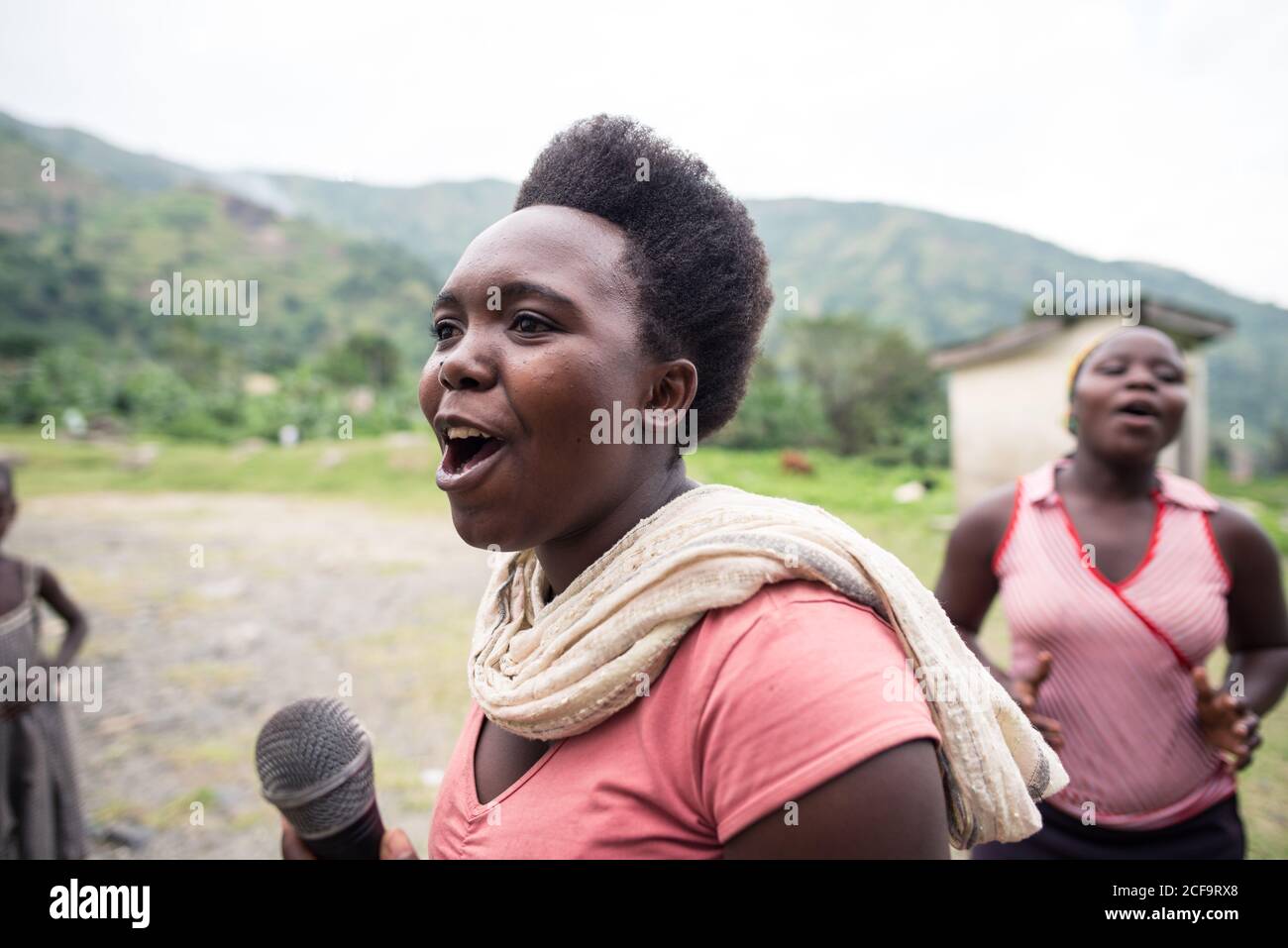 Uganda - November 26, 2016: Cheerful African female in pink shirt holding microphone and singing song while excited audience listening with interest and clapping standing against blurred green hill and small building in village Stock Photo