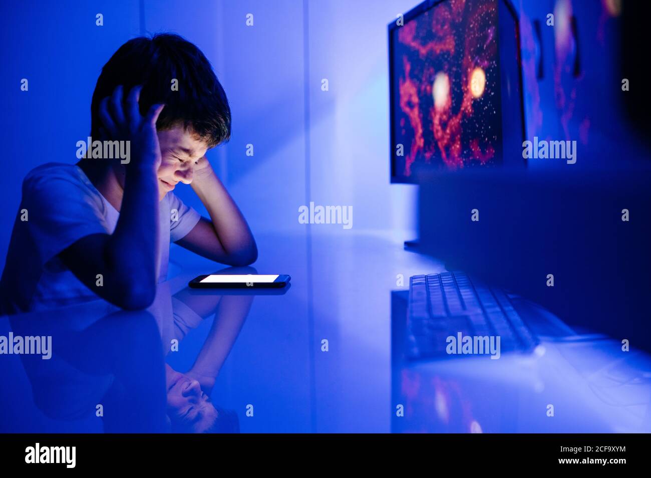 Side view of confused boy watching cellphone empty screen and touching head while sitting in room with blue light at home Stock Photo