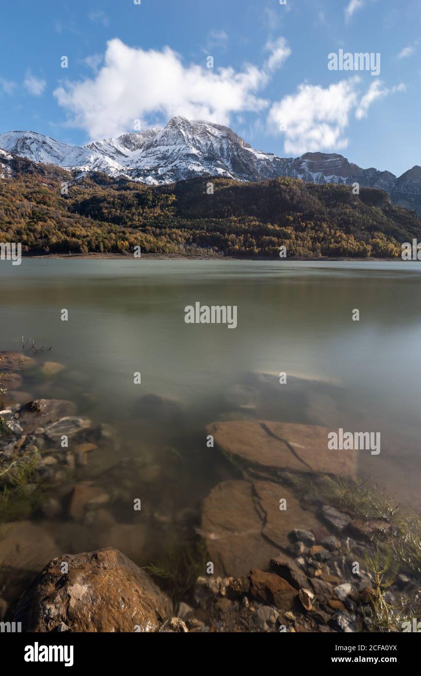 Rocks on coast of calm pond against snowy mountain ridge and cloudy sky in nature Stock Photo