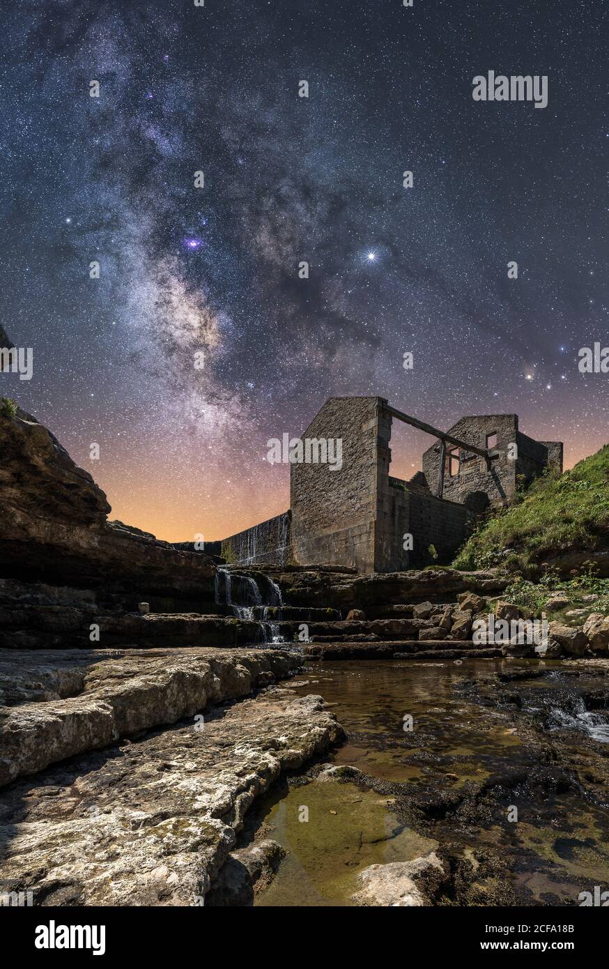 From below of ancient stone castle and small waterfall on stairs under dark sky with stars and milky way Stock Photo