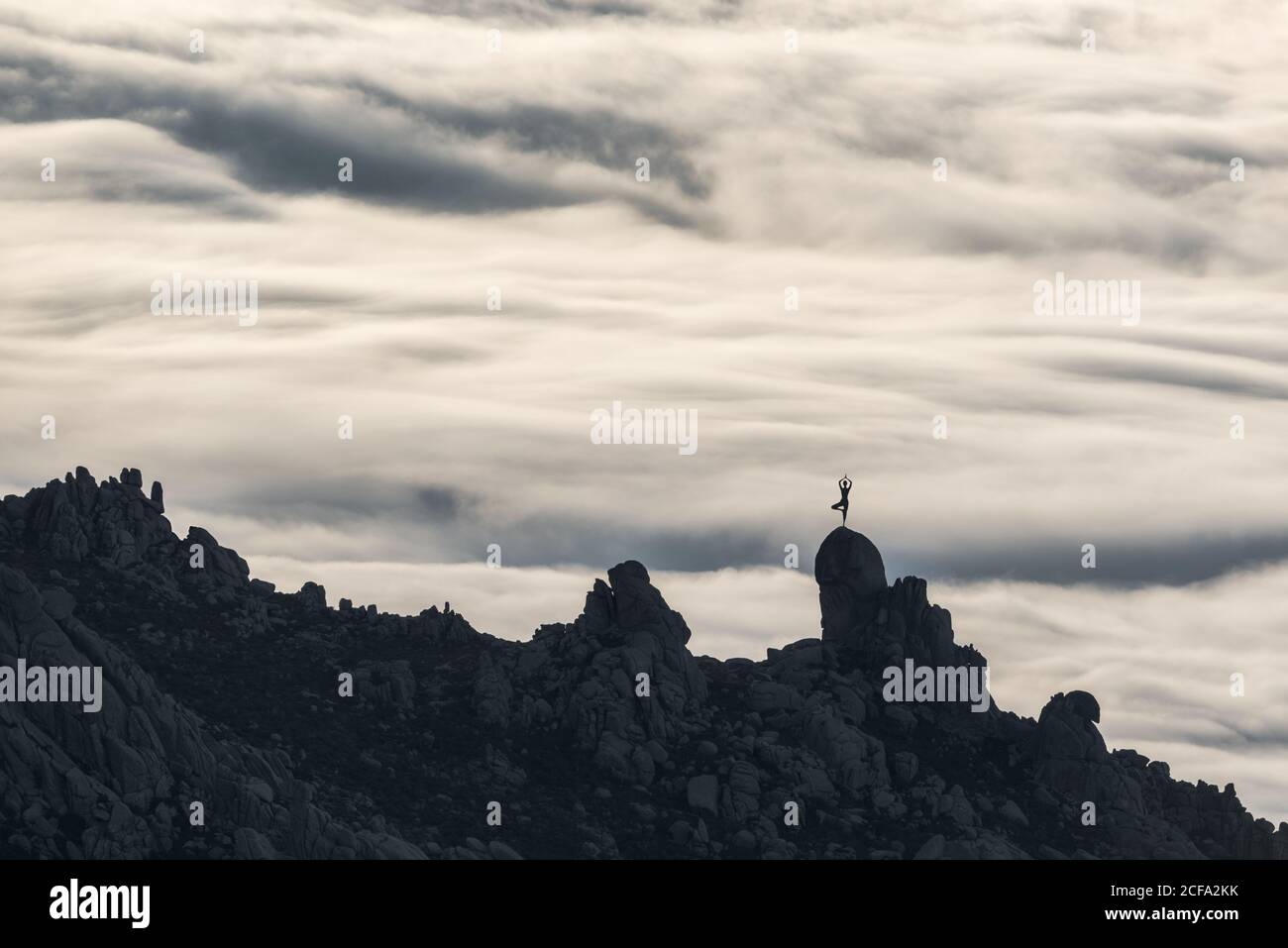 Silhouette of unrecognizable person doing yoga in graceful pose standing on peak of rough cliff with colorful clouds on background Stock Photo