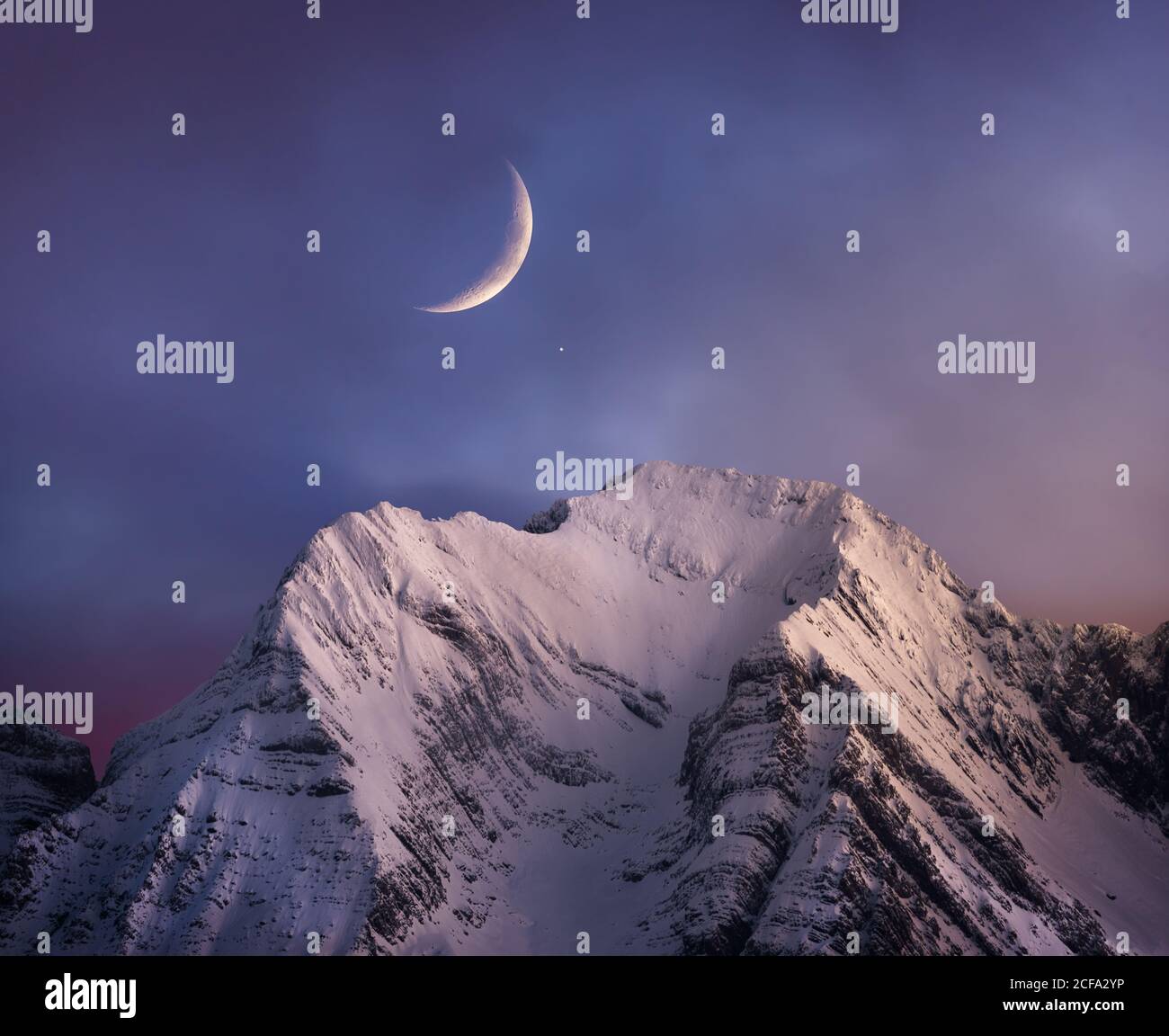 Snowy mountain range peaks under picturesque cloudy pink sky with moon at nightfall in wintertime Stock Photo