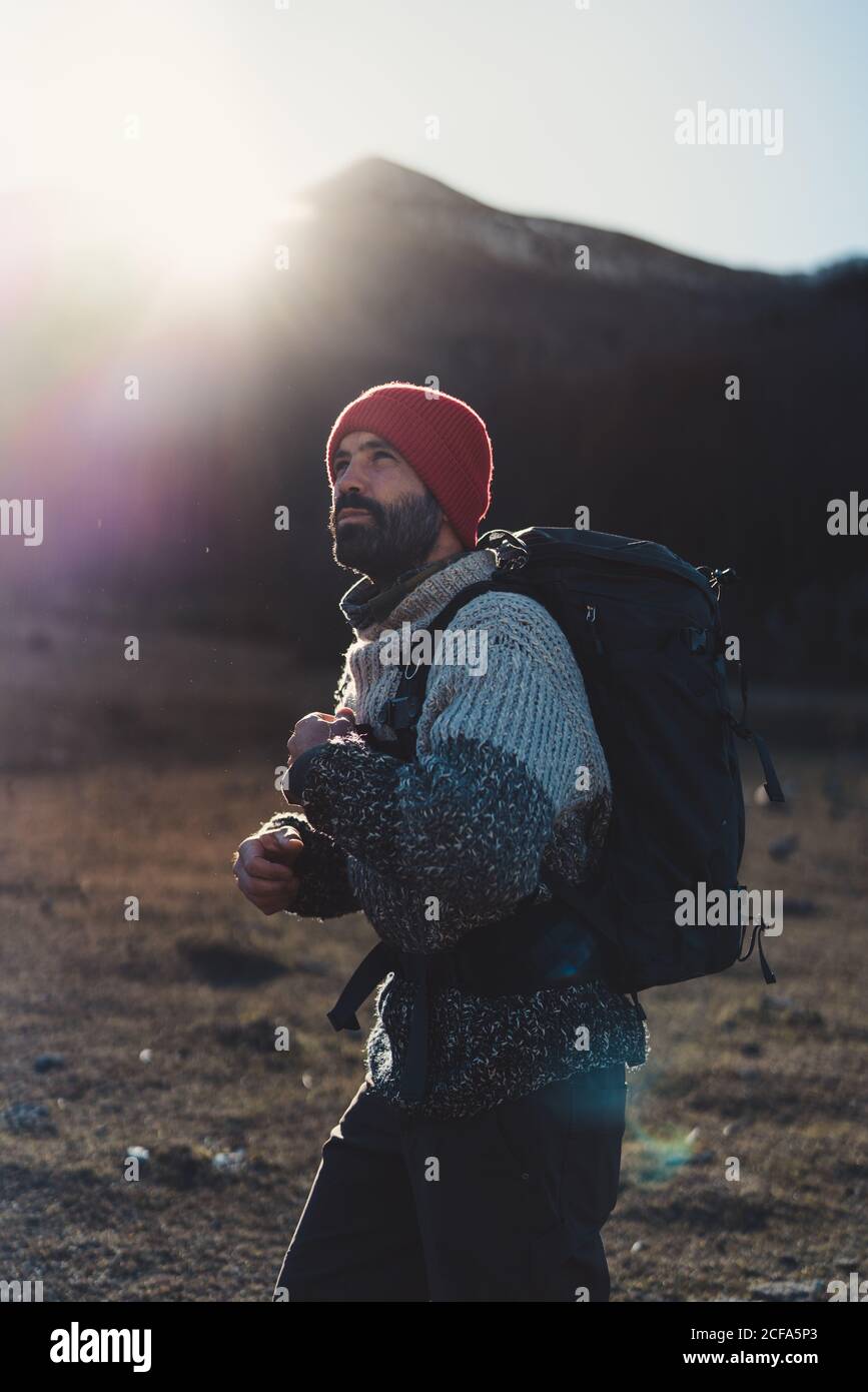 Side view of bearded male backpacker in red knitted cap and grey sweater hiking in mountains on sunny autumn day Stock Photo