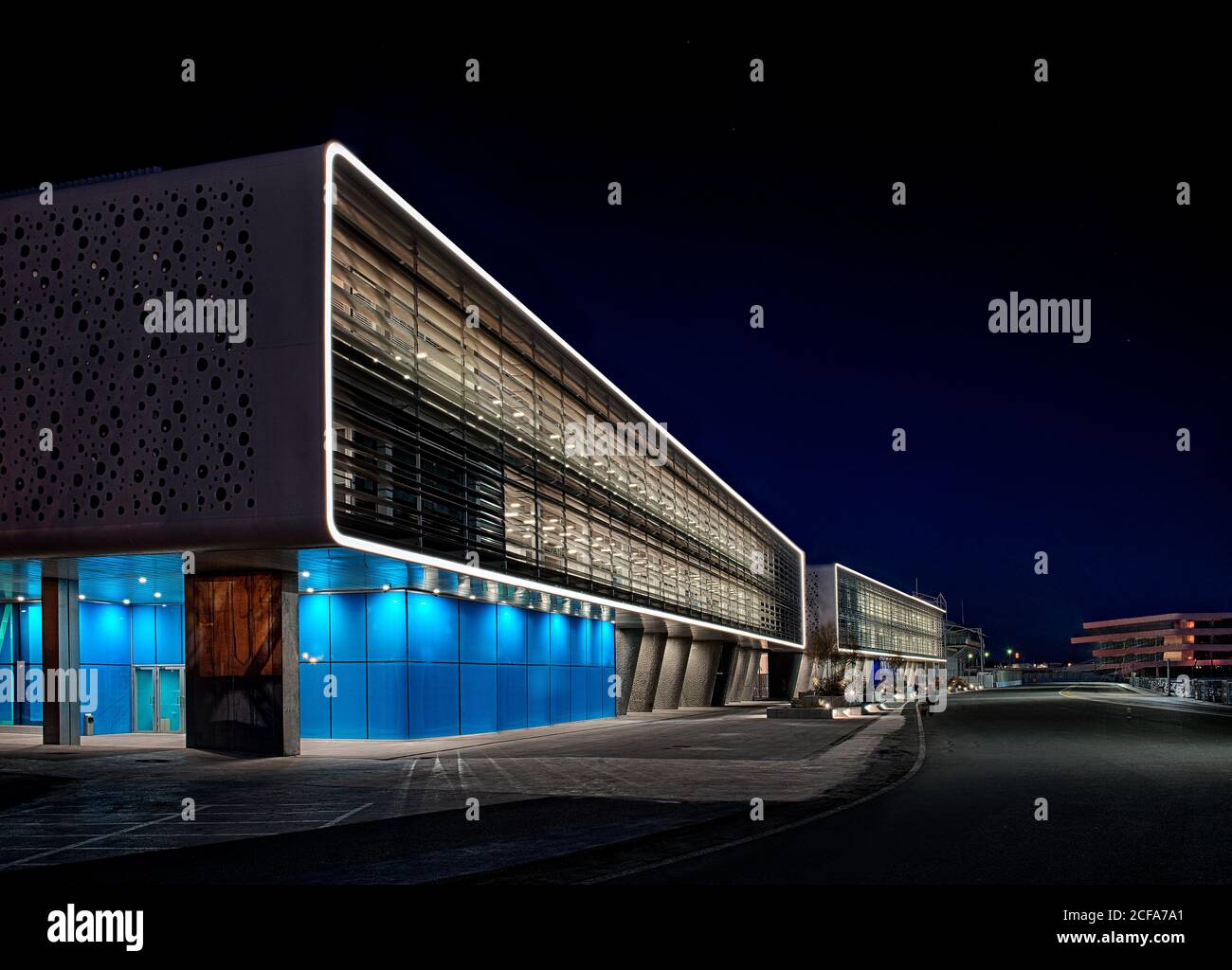 Facade of rectangular shaped building with concrete walls and vivid blue panels lighted by spot lights in composition with large striped windows against black sky at night Stock Photo