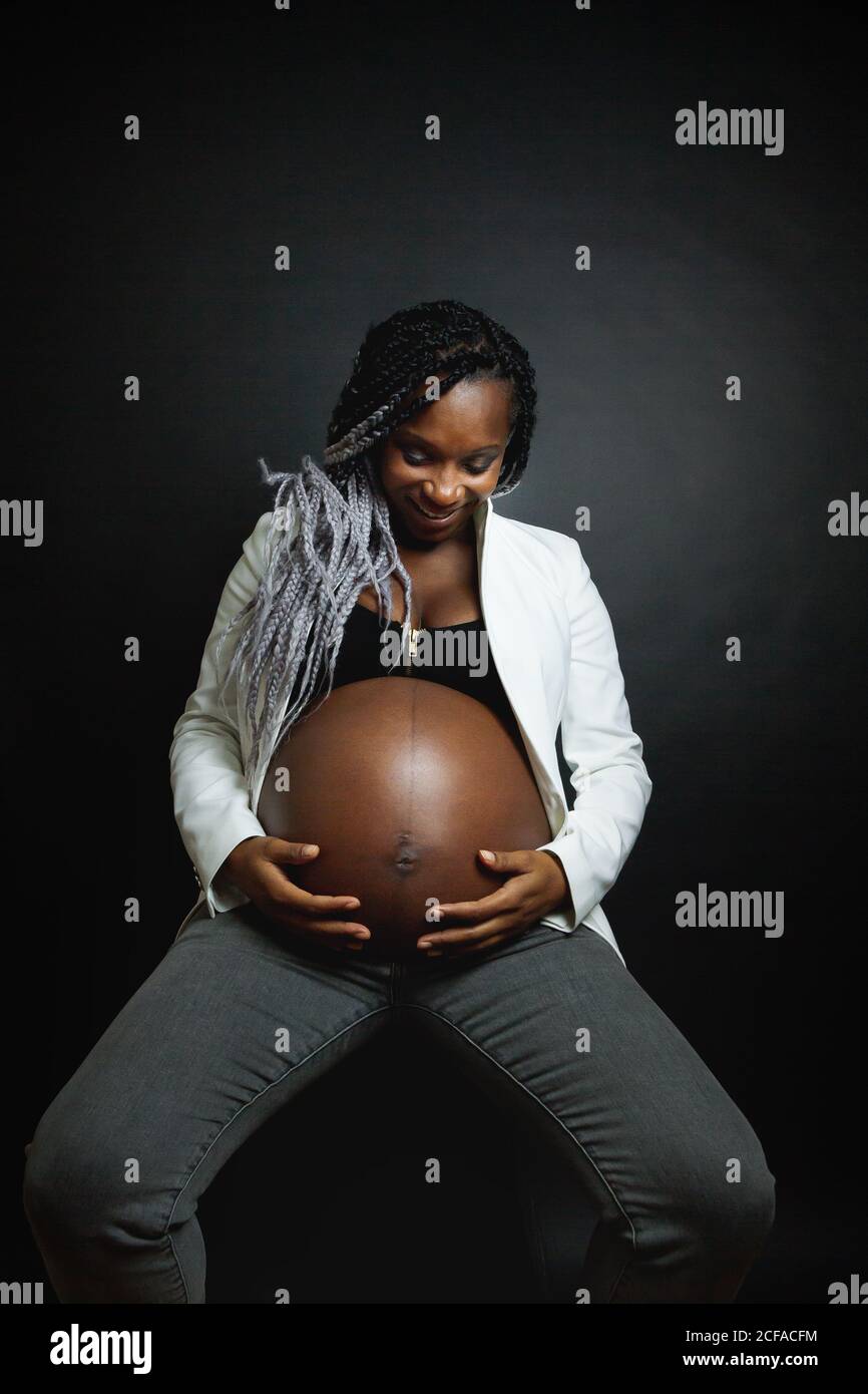 Young pregnant African American female with braids wearing white blazer and jeans stroking belly while sitting in studio against black wall Stock Photo