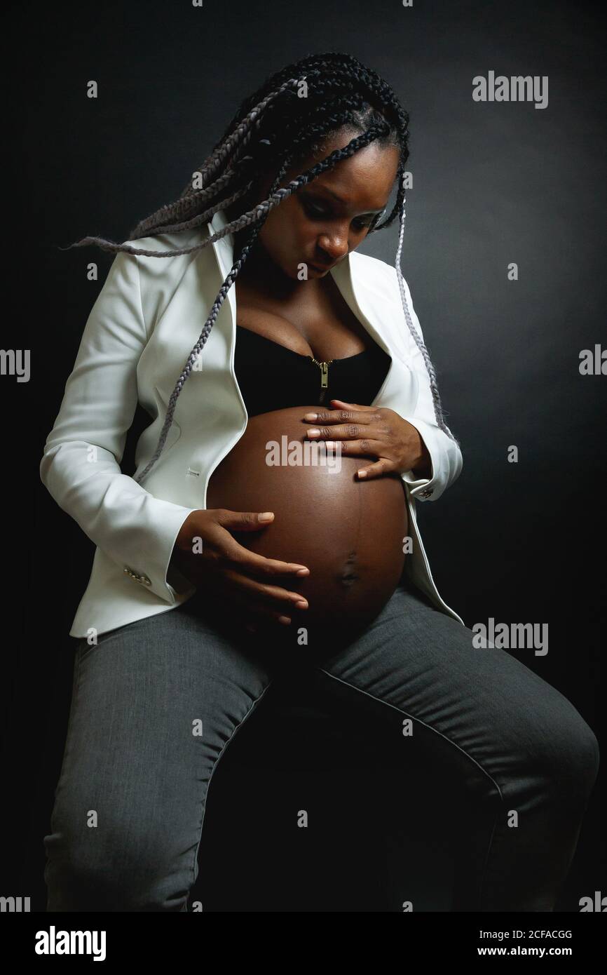 Young pregnant African American female with braids wearing white blazer and jeans stroking belly while sitting in studio against black wall Stock Photo