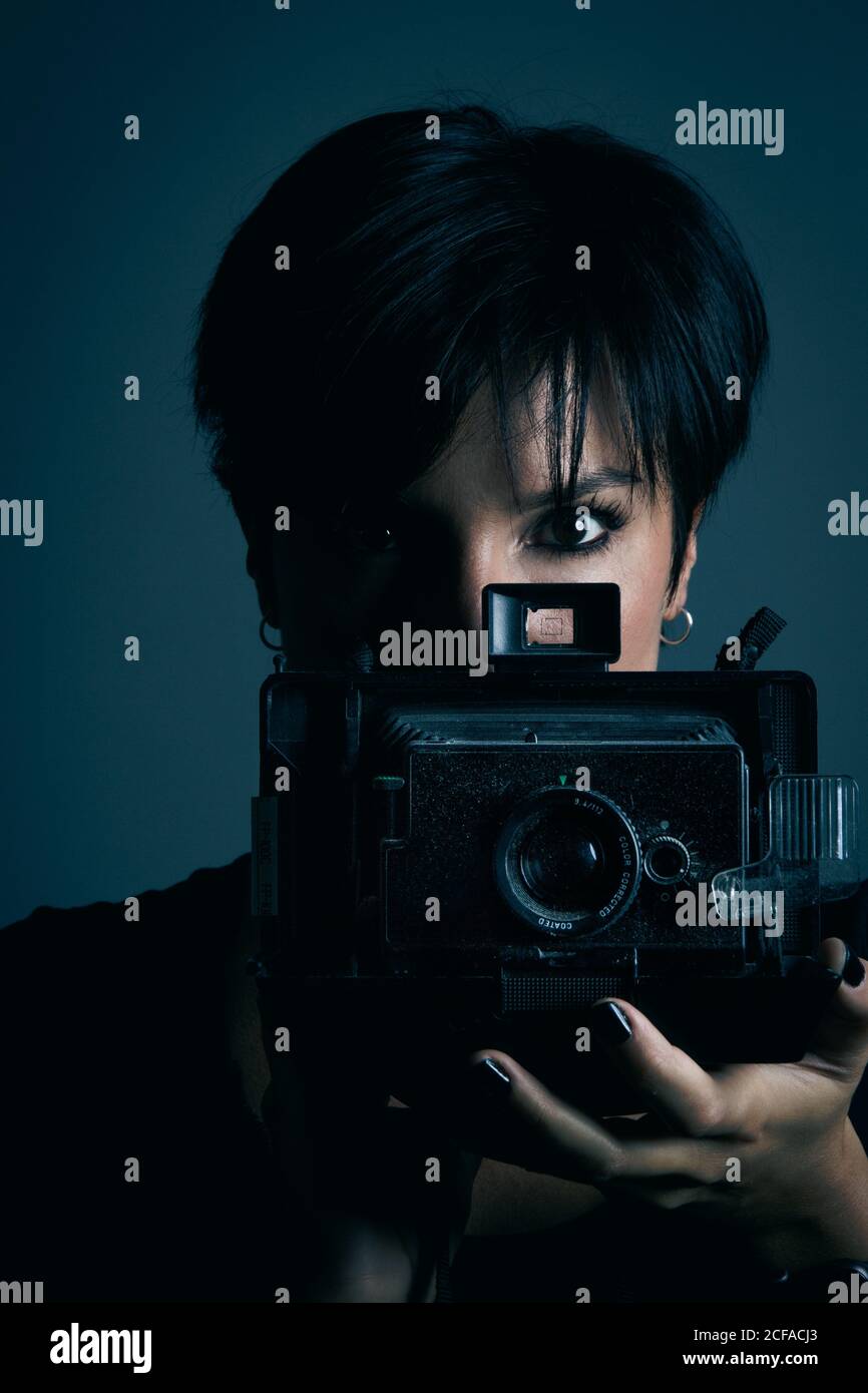 Concentrated youthful dark haired female model looking at camera and photographing with retro camera in studio on blue background Stock Photo