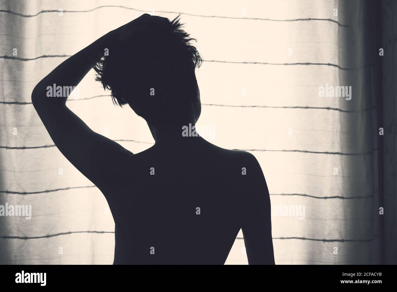 Back view of unrecognizable silhouette of female with short hair rising hand up and standing in front of window with curtain Stock Photo
