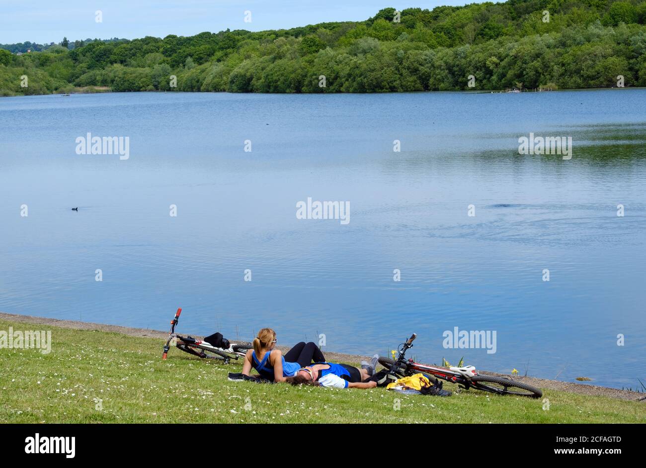Young couple lie on grass looking at water at Ruislip Lido Reservoir. Two bicycles on the ground, shoes & jackets off. Hillingdon, Northwest London. Stock Photo